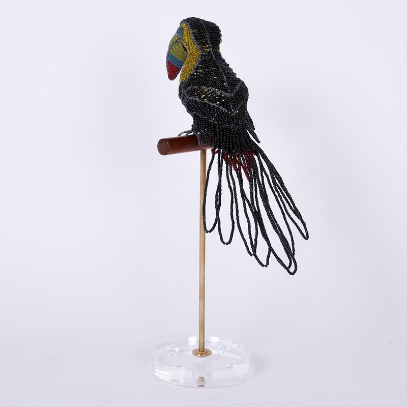 Amused and amusing bird sculpture crafted with glass beads on a metal frame and sitting on a wood perch supported by a brass rod on a Lucite base.