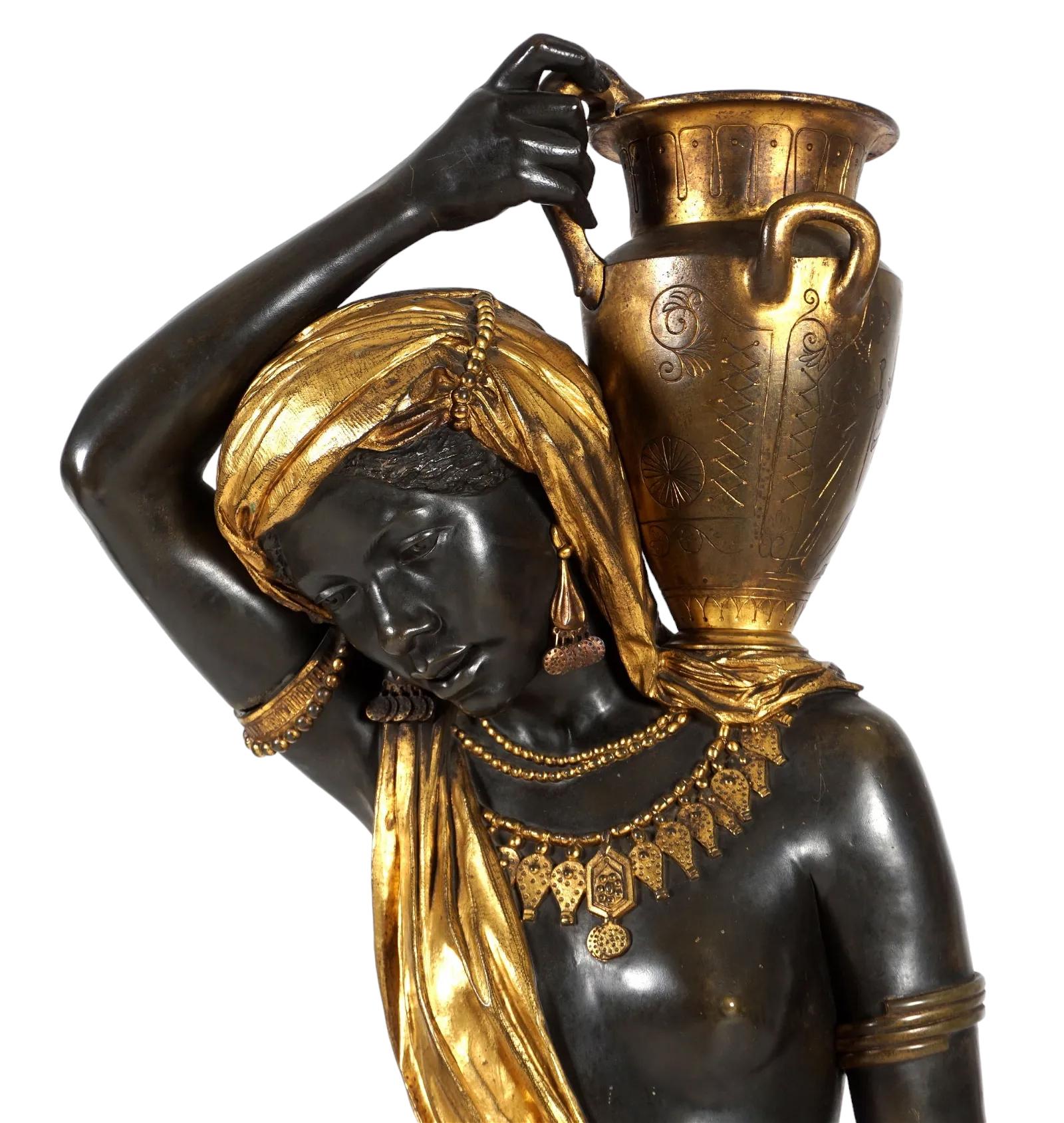 Vergoldete und patinierte Bronzestatue einer nubischen Wasserträgerin mit einem Krug auf der Schulter. Der Kontrast zwischen der tiefen Bronze und der hellen Vergoldung ist unglaublich eindrucksvoll. Die Frau trieft vor Goldschmuck und Drapierungen,