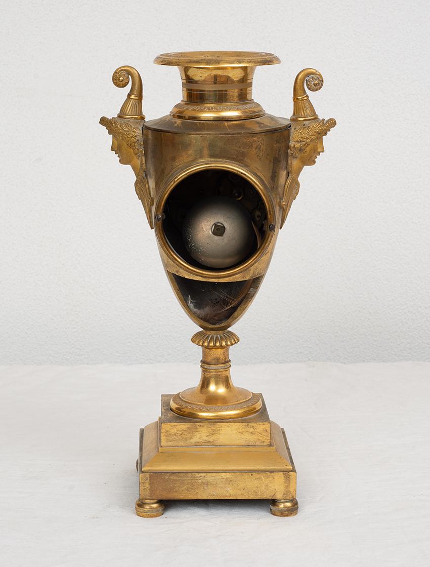 Finely chiseled gilt bronze Empire clock belonging to the early 19th century.

The vase shape is typical of its period and features a large porcelain dial surrounded by relief elements.

On the side it carries two handles depicting heads of