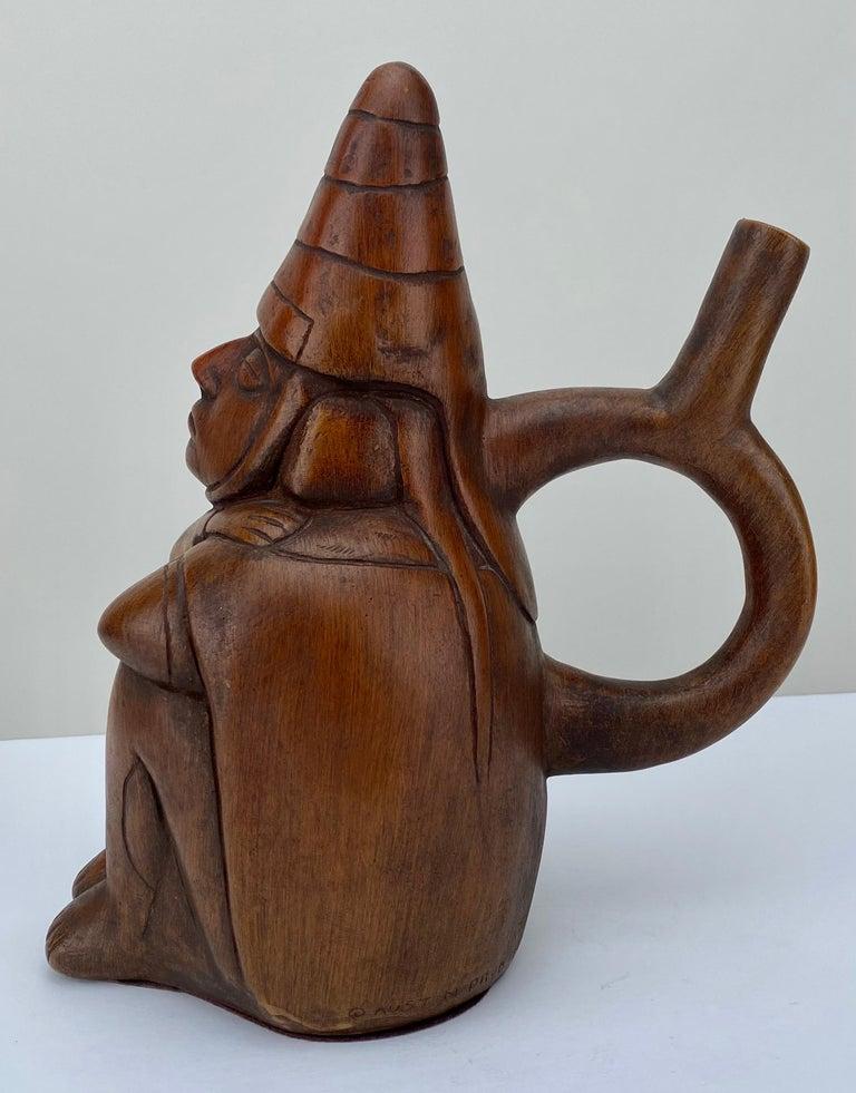 
Peruvian Figural Wood Carved Sculpture After Moche Stirrup Vessel, Dreamer:  A  stirrup vessel takes on the shape of a seated man with closed eyes, animated facial features, draped in a poncho, adorned with a conical helmet, and sporting sizable