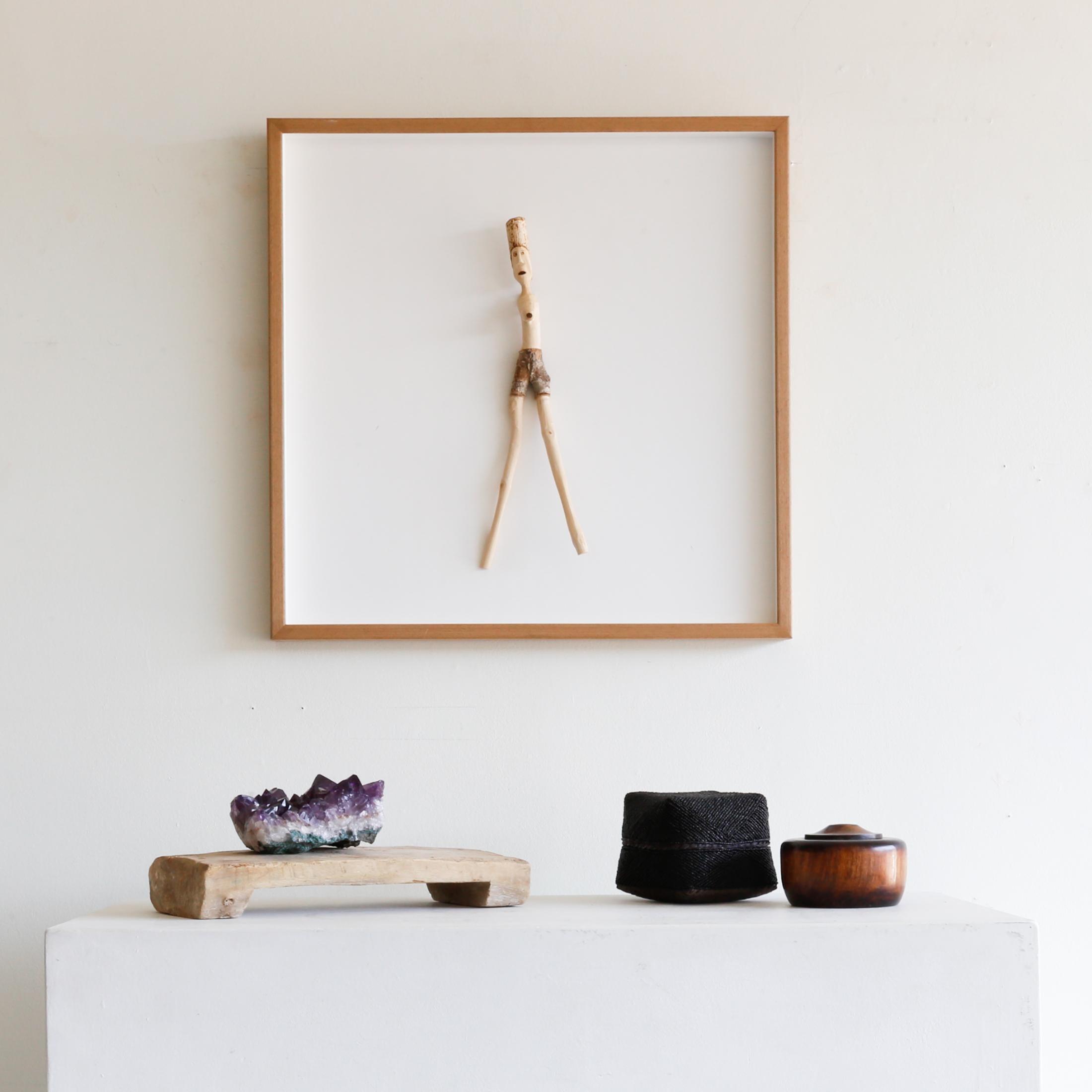 This uncanny and outlandish handmade piece features a wood figure attached to a white board in a light wood frame.