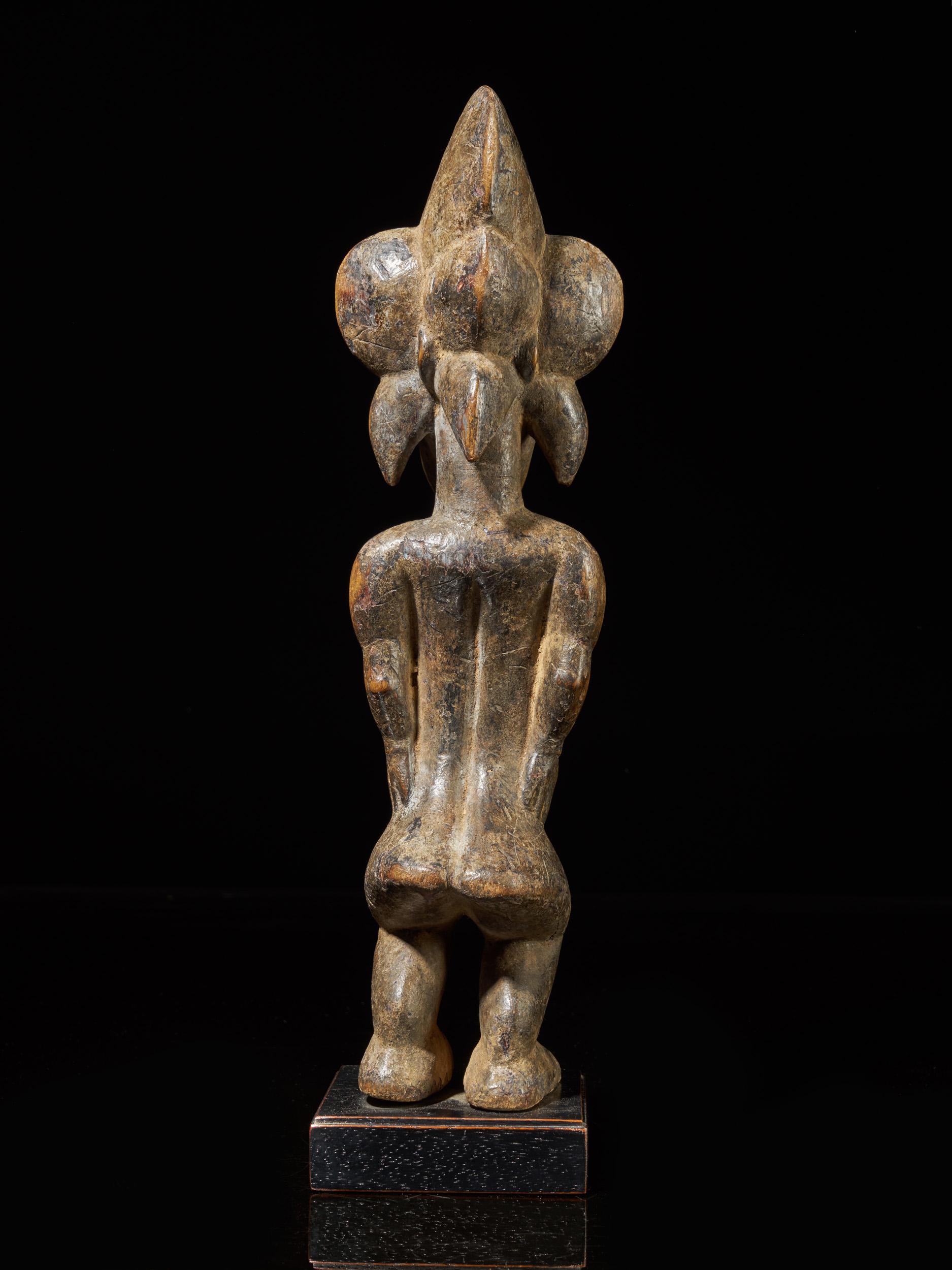 The Dolichocephalic head is on top of a robust long neck, sagittal crested headdress symbolizing mythological Birds (hornbills probably), the shoulders broad, the strong arms and legs bent, the hands to each other side of the hips, protruding belly