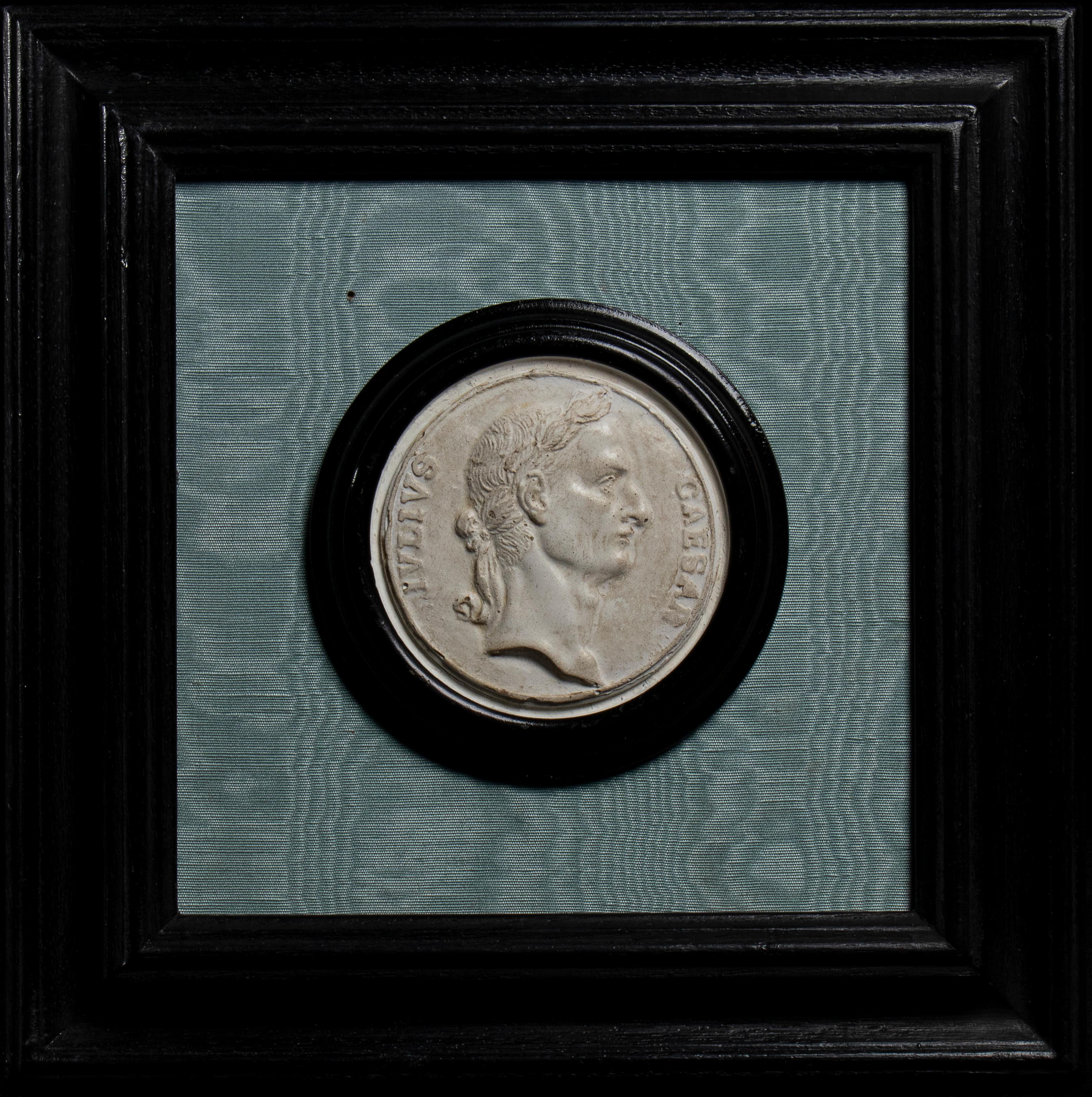 A Grand Tour collection of five plaster reliefs depicting profiles of Julius Caesar, Agrippina, and the Roman emperors Vitellius, Otho, and Vespasian. Mounted in black frames against a light gray-blue satin background. Each profile is enclosed in a