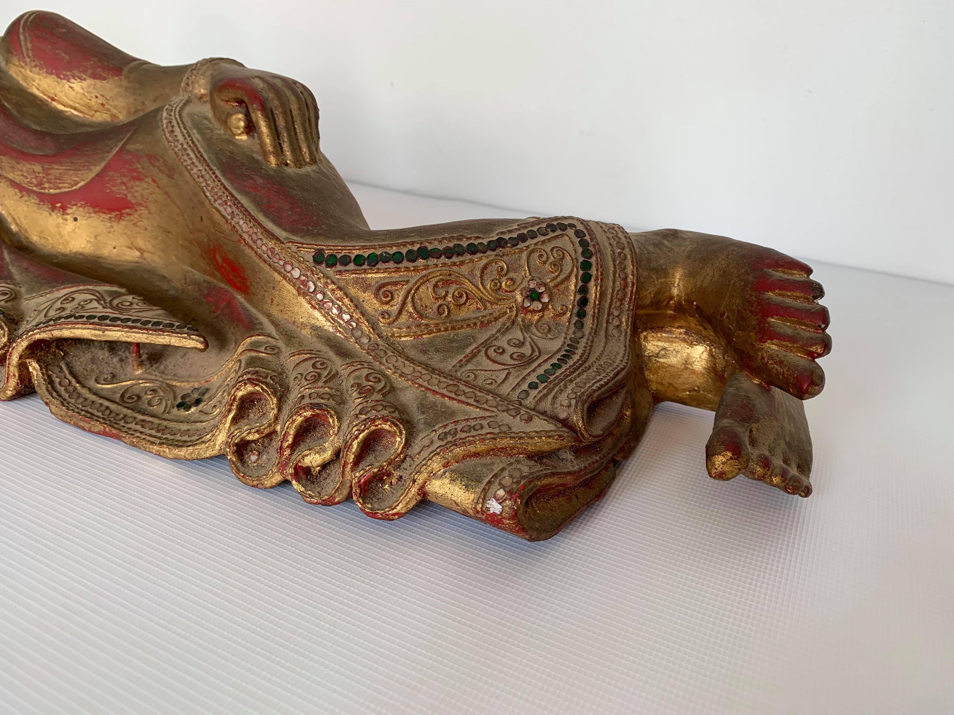 This is a mid-century reclining Buddha from Southeast Asia, most likely originating in Cambodia. It is carved from wood, gilded and painted, and ornamented with clear and green embellishments. It has gracious lines, appropriate for displaying on a