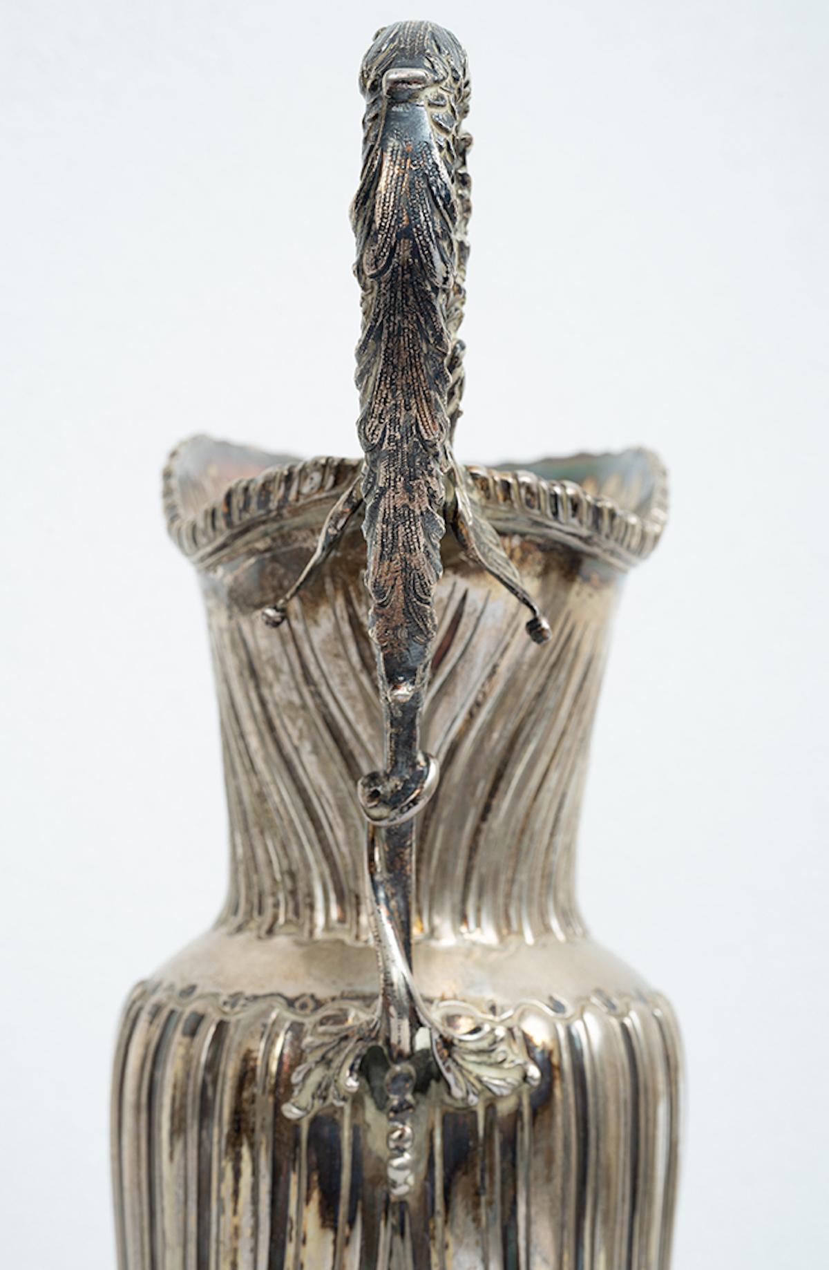 Antique Neapolitan silver pourer belonging to the early 20th century.

The central sinusoidal-shaped body is embellished with a pod interspersed with a winged dolphin-shaped handle.

The base is oval in shape embellished with dense chiseling on the