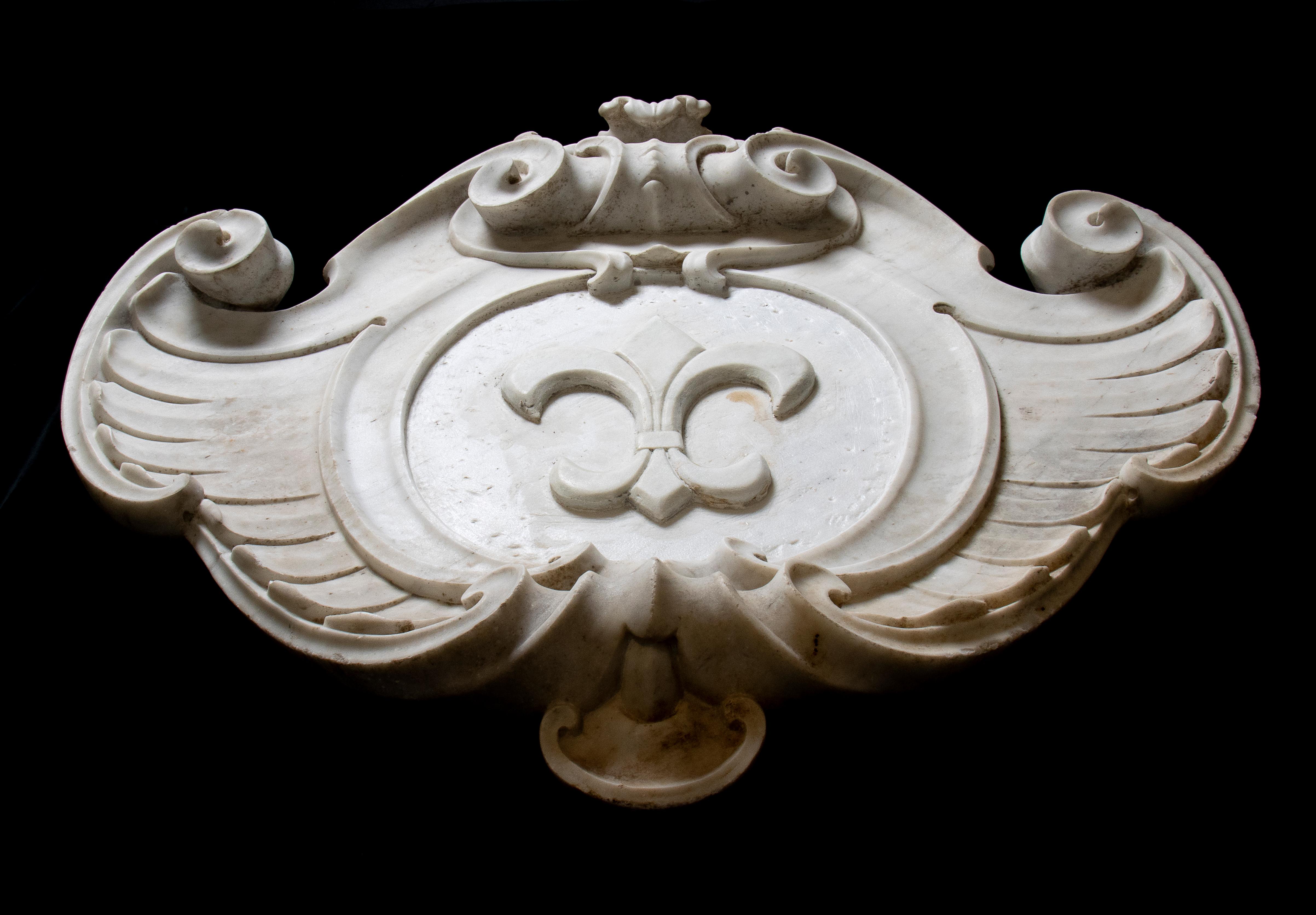 Ein sehr interessantes Wappen aus weißem Marmor mit symmetrischer Struktur und doppeltem Rollendekor sowie Akanthusblattschnitzereien. Mit zentraler Darstellung einer Lilienblüte im Wappen.
Die Fleur-de-Lis, auch Fleur-de-Lys genannt, ist ein