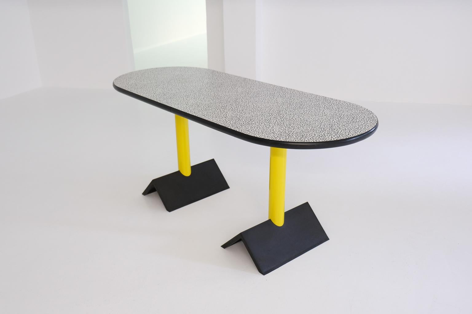 custom made oval dining table or desk, or console table in memphis milano style. the oval top is laminated with the classic memphis squiggle pattern and framed by a semicircular rubber edge. the yellow metal legs are set in cast-iron feet shaped