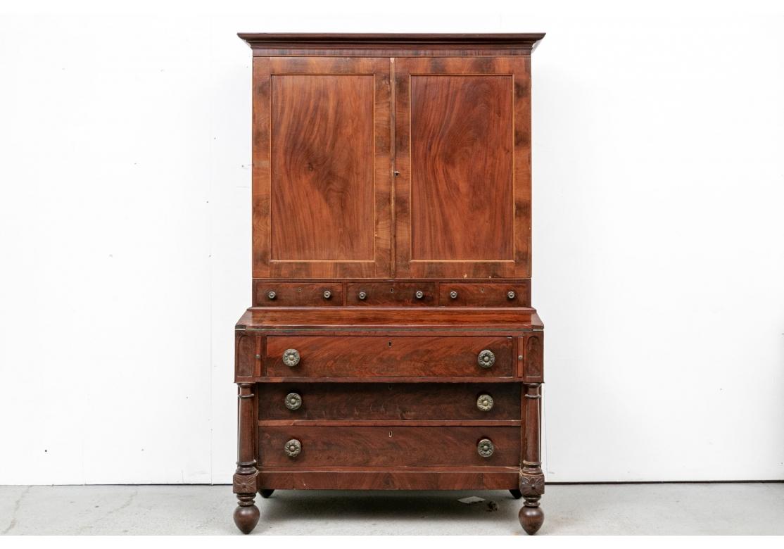 Figured wood veneer, the top with a carved cornice over double doors. They open to two shelves over drawers and cubby holes. A middle frieze with three drawers (lacking a key) over the lower projecting case with slant front with green felt writing