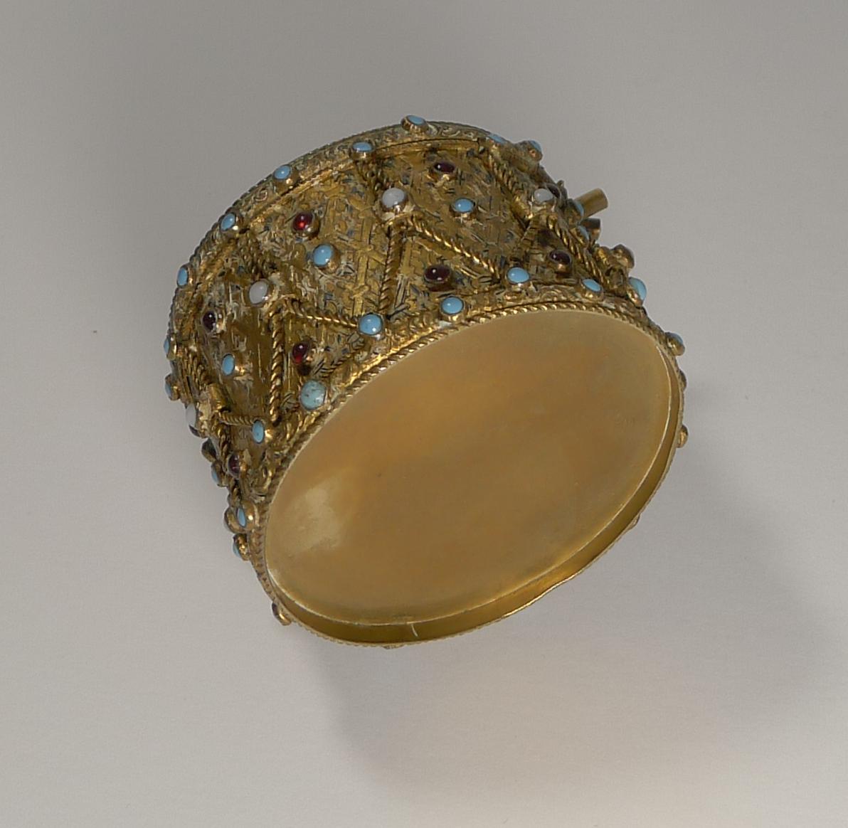 Eine höchst ungewöhnliche Schatulle aus dem späten 19. Jahrhundert in Form einer Trommel aus vergoldetem Messing mit einer Auswahl an unechten Cabochon-Türkisen, Opalen und Granaten.

Die Griffe der beiden Trommelstöcke werden über den Rand der