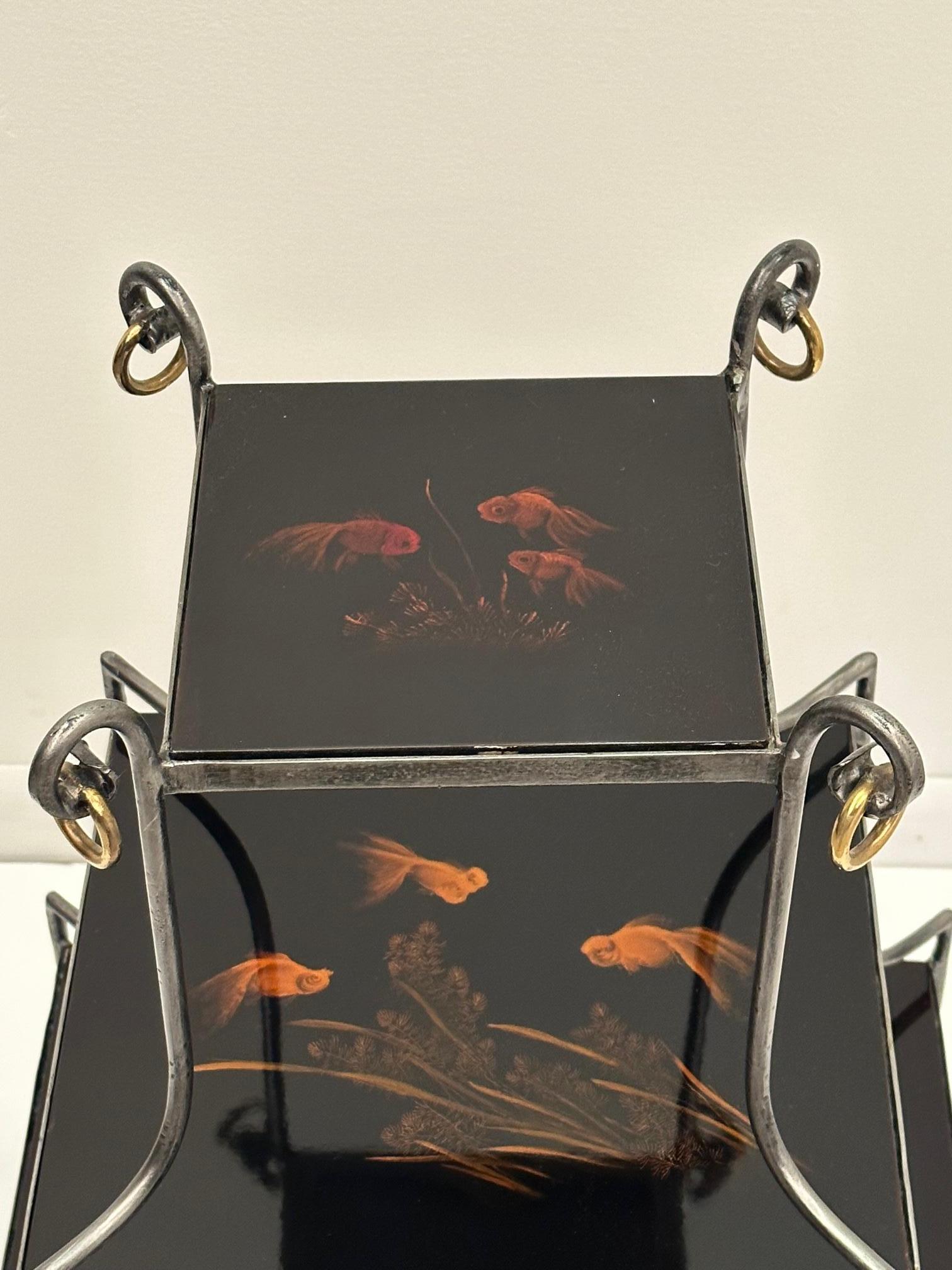 Wunderschön gestalteter, ungewöhnlicher 3-stufiger Beistelltisch mit einer Form aus brüniertem Stahl und Messing und schönen, schwarz lackierten, abgestuften Tischflächen, die mit Goldfischen verziert sind. Schöne Messingringe an der Spitze.
Obere