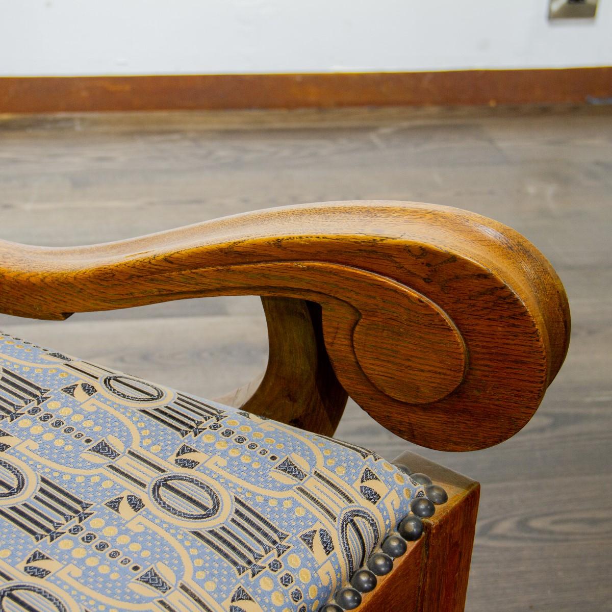 A Scandinavian oak art nouveau child sized chair with chunky legs and organic scrolling arms. The back rest of the chair has oak rivets down the side of the frame and finials to the top. The chair has been upholstered in a Backhausen art deco