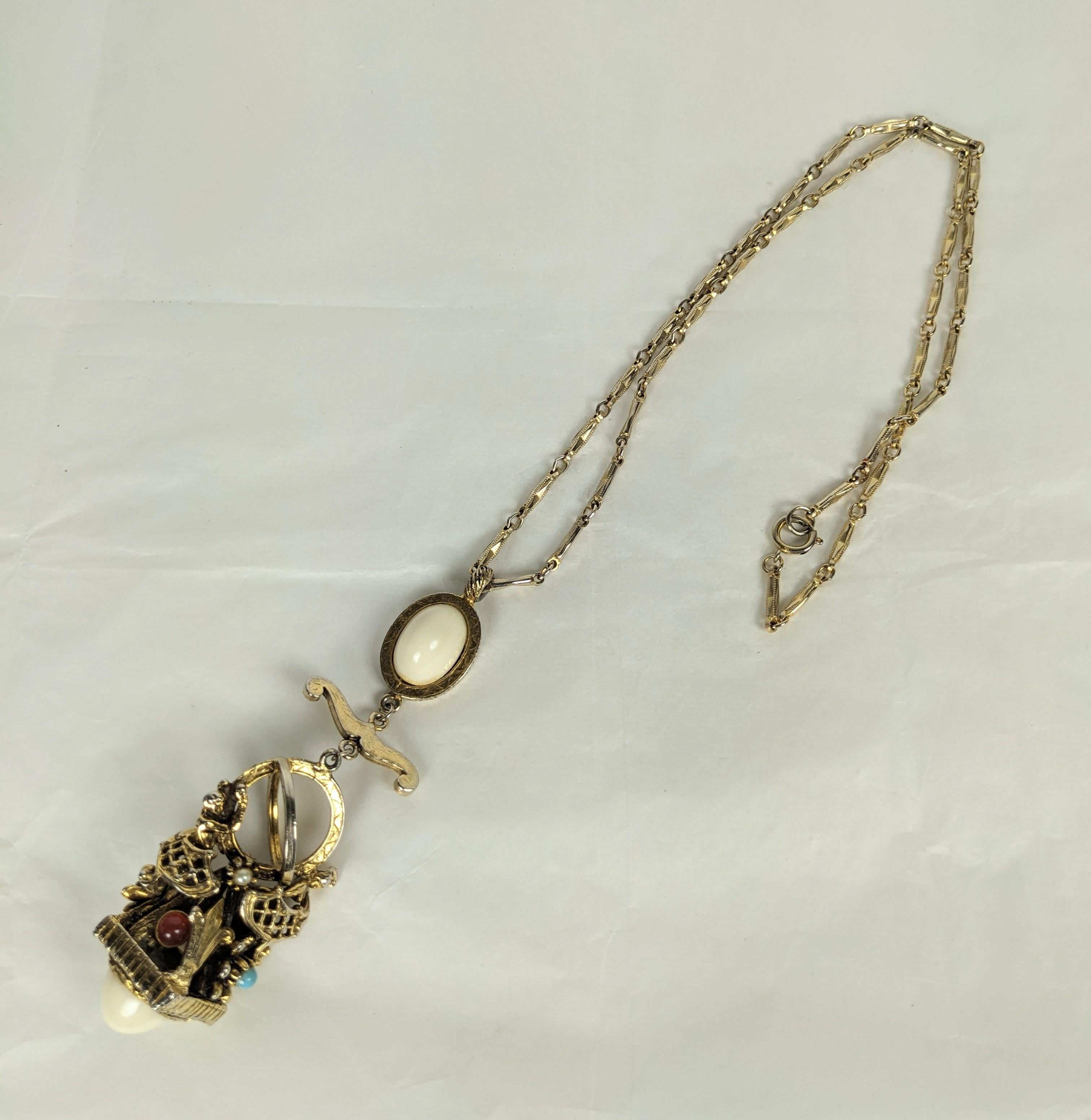 Ciondolo a forma di ciondolo fantasy degli anni '60 raffigurante due figure che sorreggono un mappamondo con fleur de lis ed elementi floreali decorativi. I cabochon in vetro colorato sono punteggiati da cabochon in finto avorio alla base e in cima.