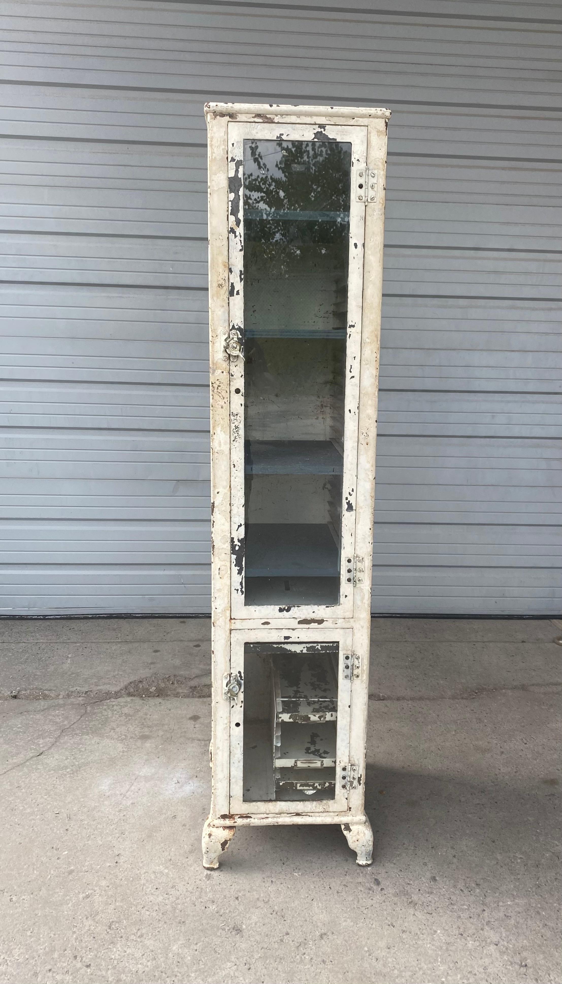 Unusual industrial medical / specimen cabinet, steel and glass. Great size, scale, proportion. Tall narrow metal cabinet, upper glass door with shelves, lower glass door will small pull out drawers. Retains original white paint, as well as turning