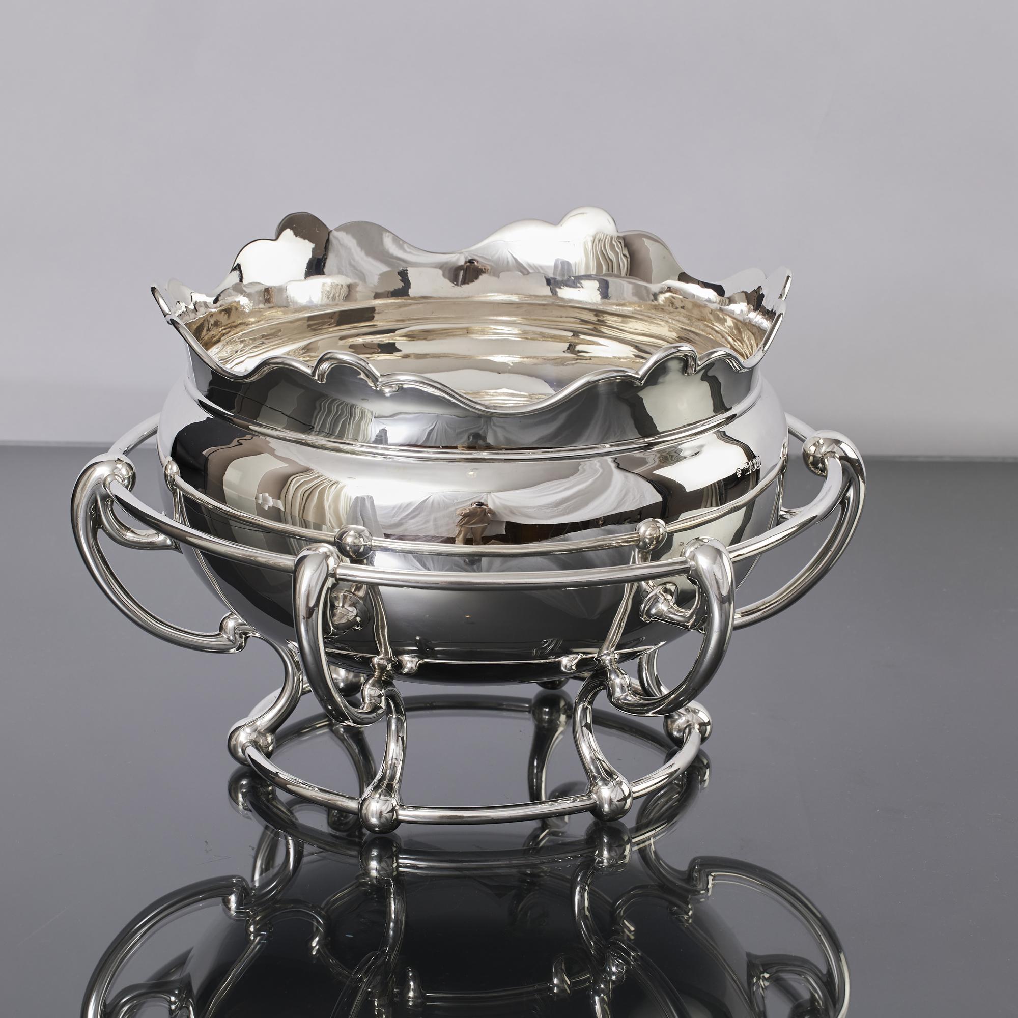 Gran cuenco antiguo de plata de ley, con borde ondulado y alojado en un marco abierto construido con raíles tubulares y soportes perfilados con volutas.
Aunque el riel del marco es una parte decorativa del diseño, podría utilizarse para colgar tazas