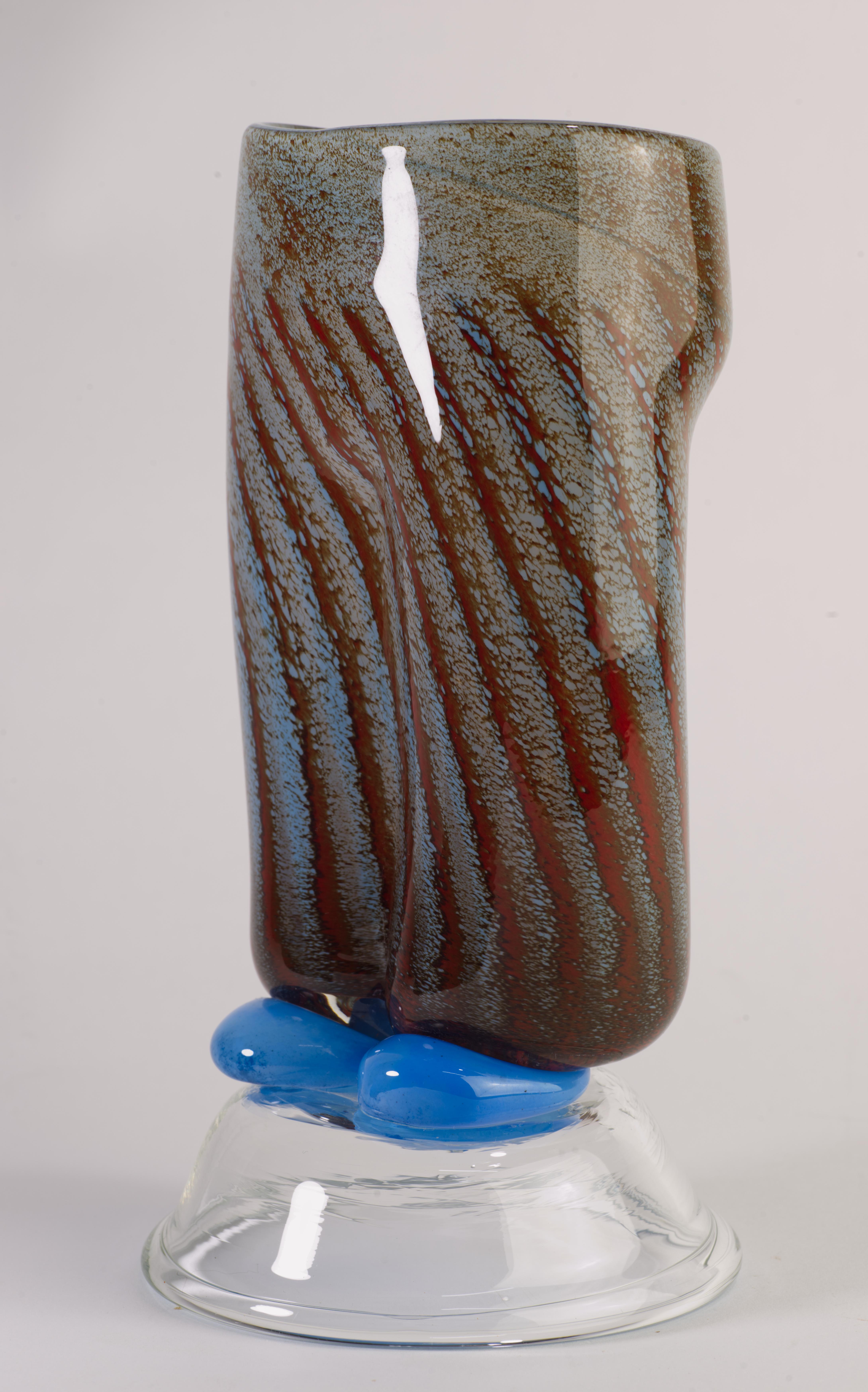 Un vase en verre d'art inhabituel et ludique a été créé en forme de bas de corps avec un pantalon bleu tacheté avec des rayures rouge foncé descendant sur des chaussures bleu clair ; les chaussures et le piédestal rond en verre transparent forment