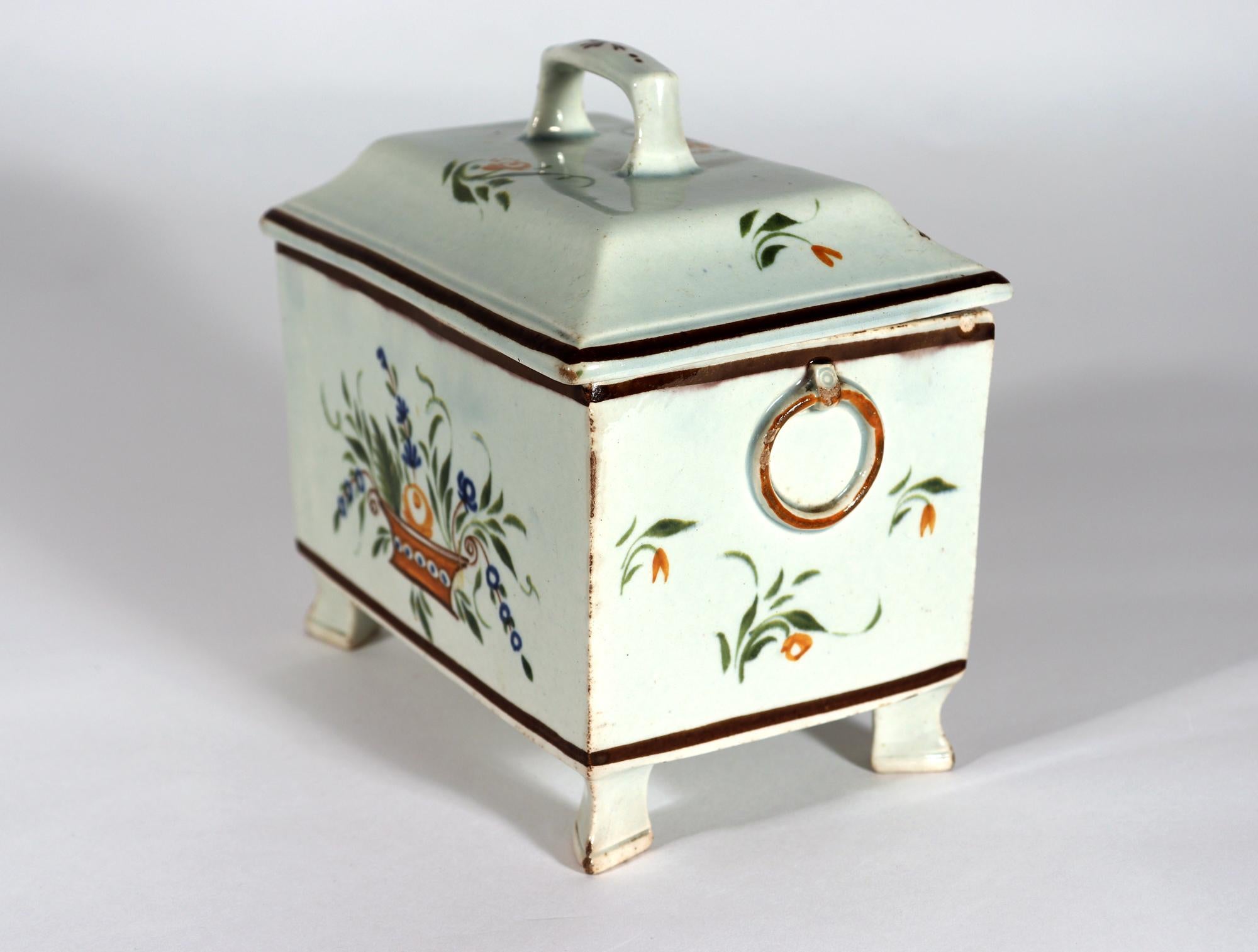 Boîte à thé Botanique couverte en poterie de Pearlware Prattware,
Poterie cambrienne, Swansea
Circa 1800-20

La boîte à thé à pied de forme rectangulaire en poterie perlée est peinte aux couleurs de Pratt (bleu, vert et orange) sur chaque côté