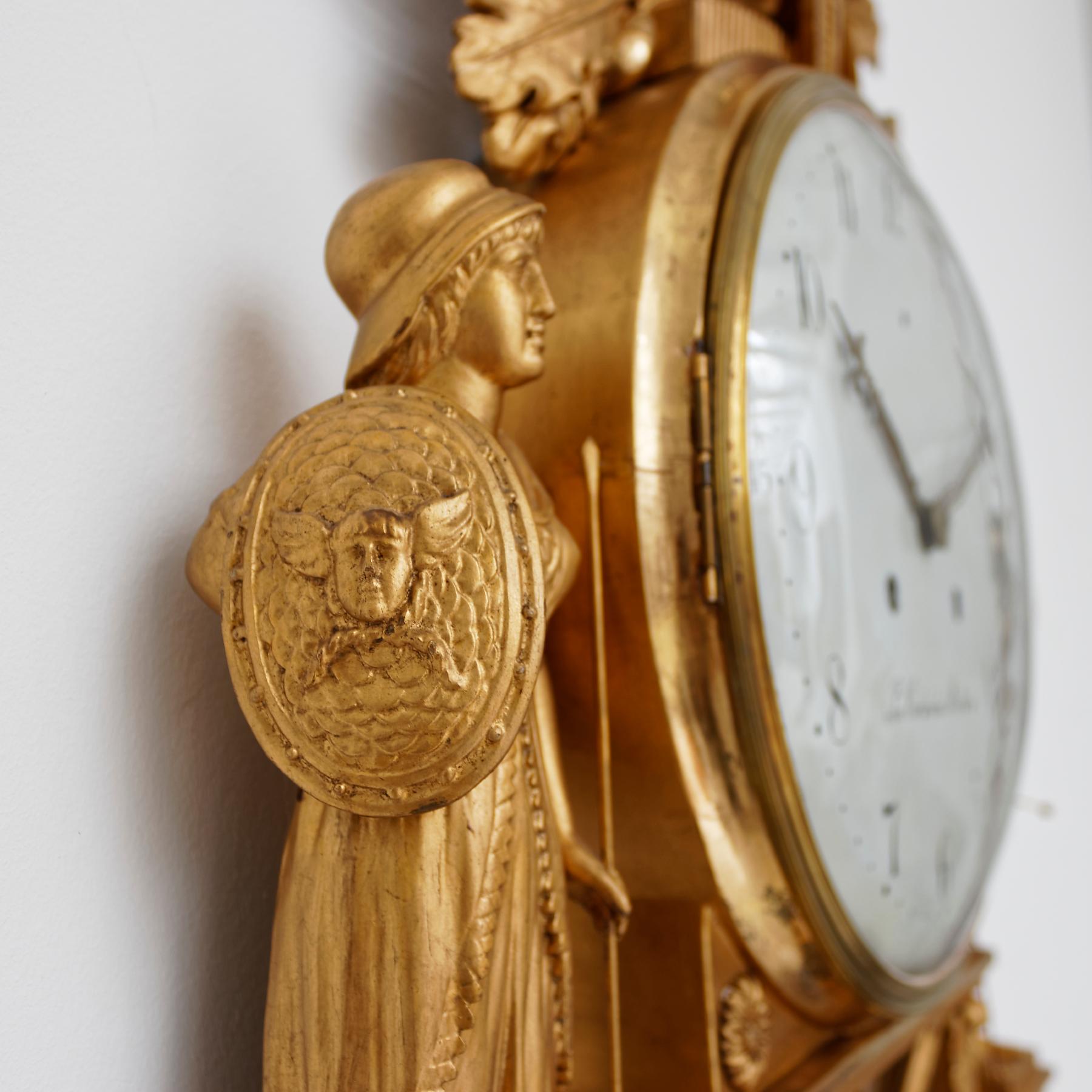 An unusual Swedish Empire giltwood striking wall clock from the early 19th century. The white painted Arabic dial signed by the clockmaker ‘Jon. Cederlund Stockholm’ (Jonas Cederlund 1768-1857). The clock is surmounted by a military trophy with a