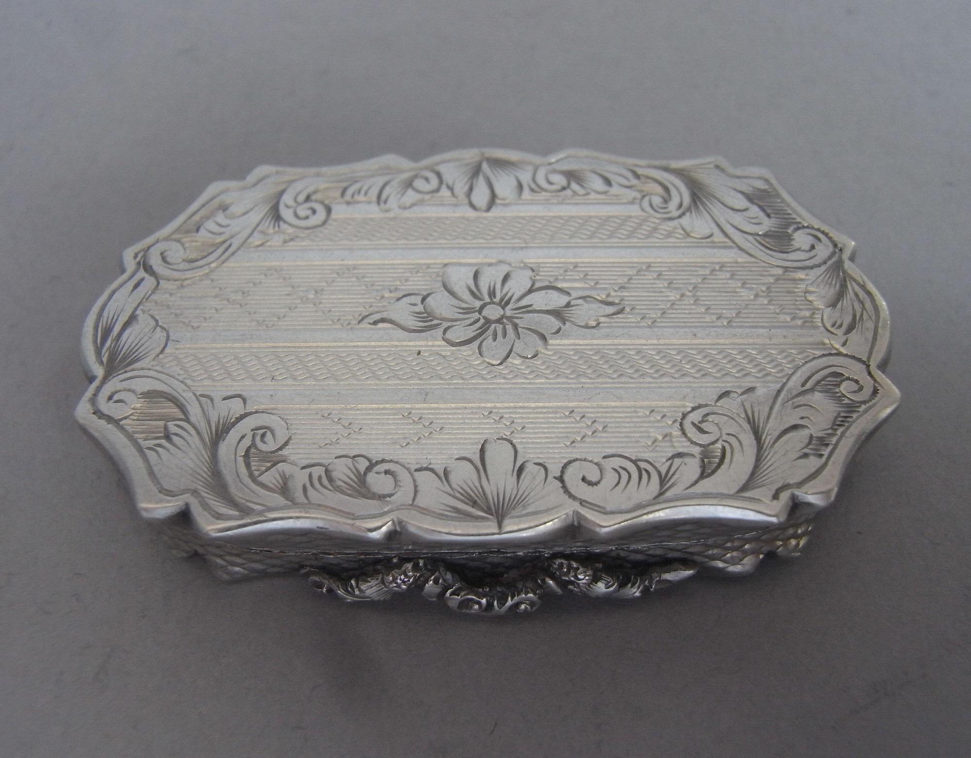 An unusual Vinaigrette made in Birmingham in 1848 by Francis Clarke.

The Vinaigrette is modelled in an unusual shaped rectangular form. The cover is engraved with an outer border of feathery scrolls enclosing a panel of various horizontal bands of