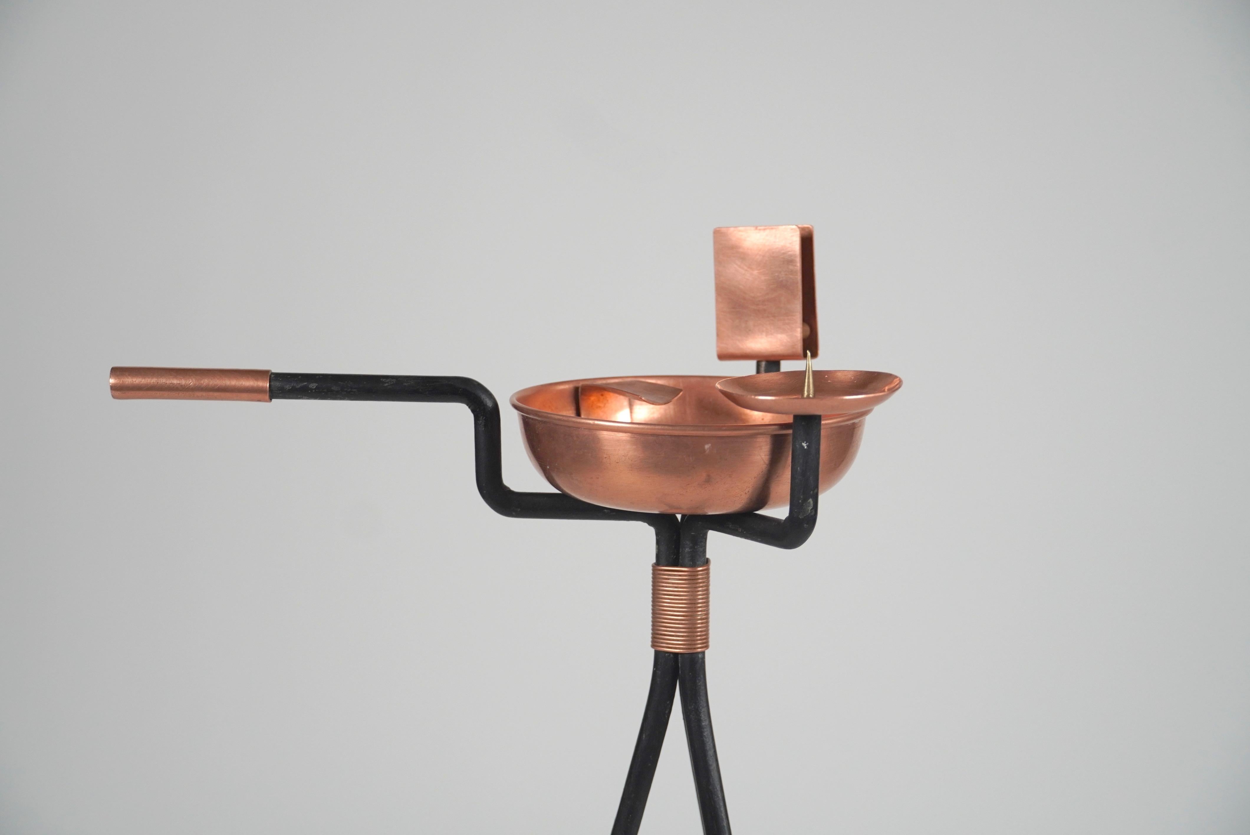 An unusual design in a cigar floor stand ashtray, created out of copper, brass, with a three legged iron base wrapped in copper wire which has a candle holder to relight the cigar and a match box holder to relight the candle. The handle allows the