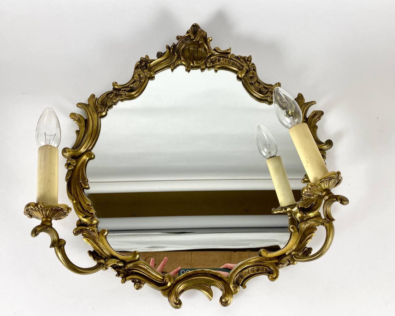 Baroque-style circa 20th century bronze colored brass two-light girandole mirror.

The oval shaped mirror with pierced scrolled acanthus cresting and flanked by pilasters and floral festoons.

The frame structure is made of brass. Very detailed