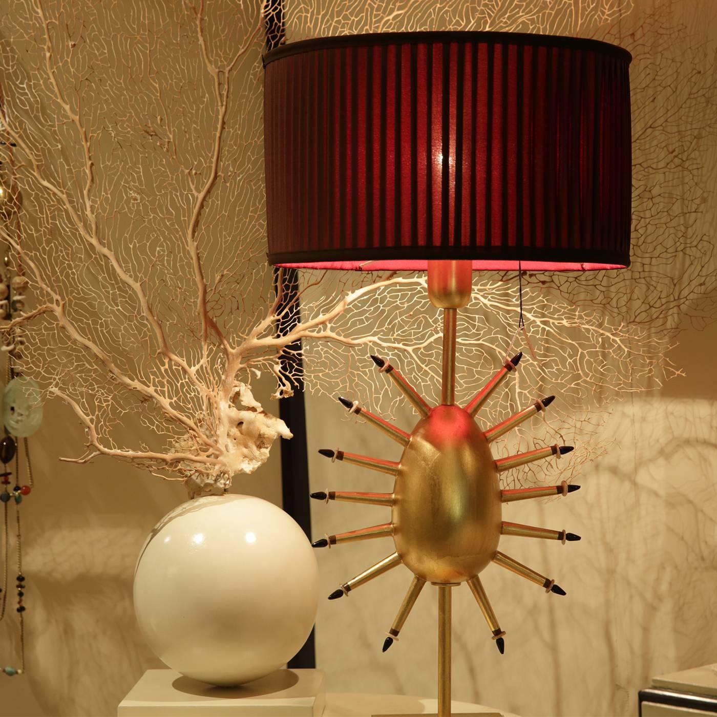 This radiant wooden table lamp in a golden finish by Bottiglieri sits with easy brilliance under a moody crimson shade of hand-sewn silk lining. The central form of the egg and its extending rays of precious stones express transformation.