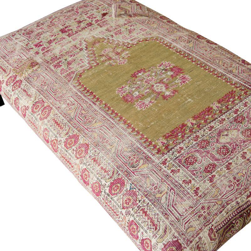 A beautiful custom-made ottoman or coffee table with a simple black metal base. The ottoman is made using a vintage rug, complete with beautiful colors, designs, and patina. The top is a mix of mostly off-white, red, yellow, and black. The base is