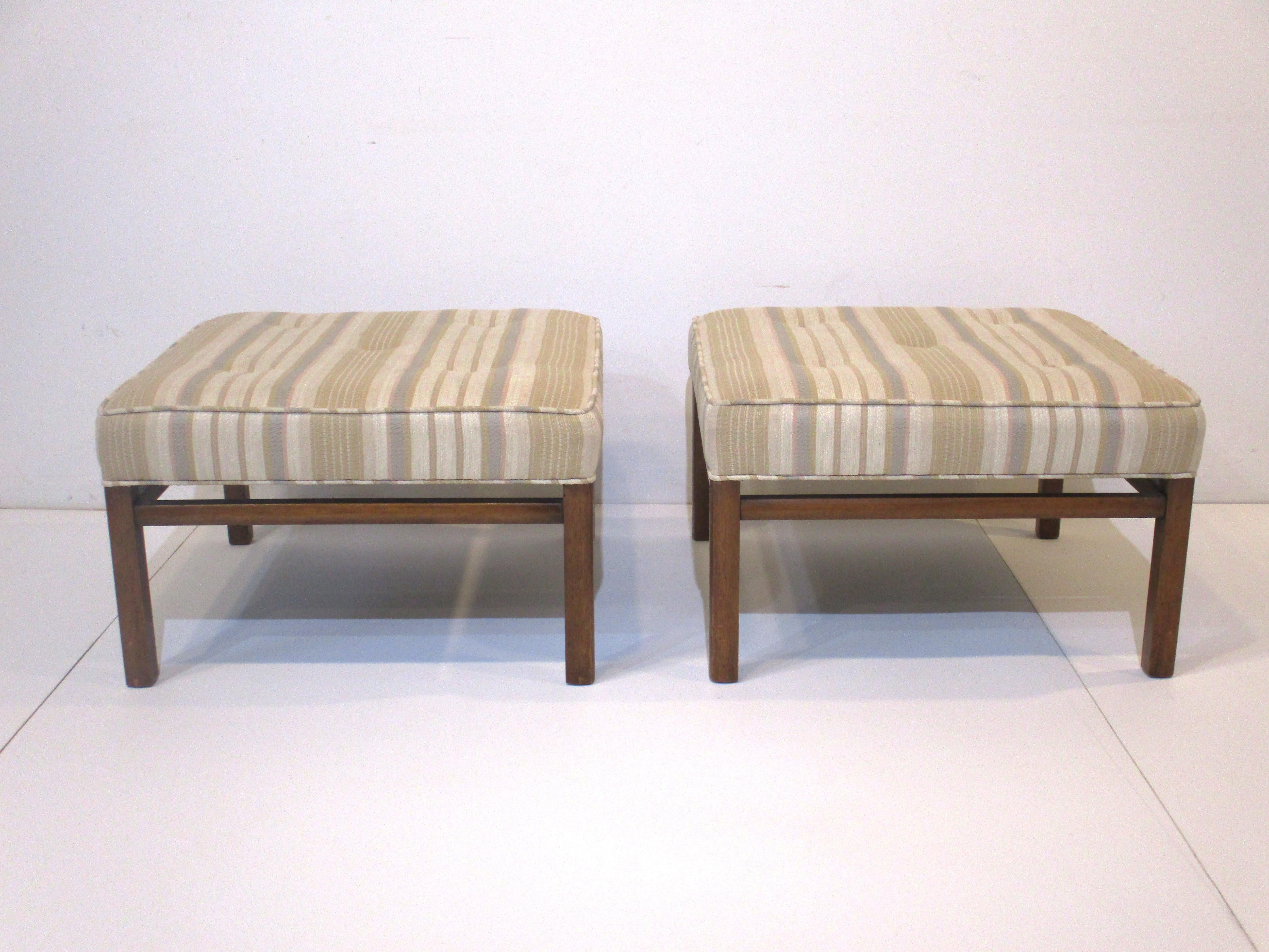A pair of upholstered ottomans or stools with dark toned mahogany frames and legs . The top stripes in the fabric are creams, taupes , gray and tans with buttons giving them a tight look and feel in the manner of Harvey Probber.