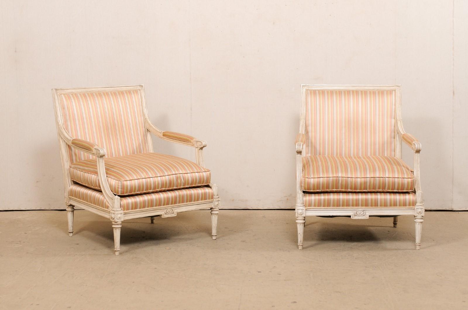 A vintage pair of American carved-wood and upholstered armchairs. This pair of French style fauteuils have a Louis XVI inspired design, with square-shaped upholstered backs framed within carved molding, arms with manchette padding that terminate