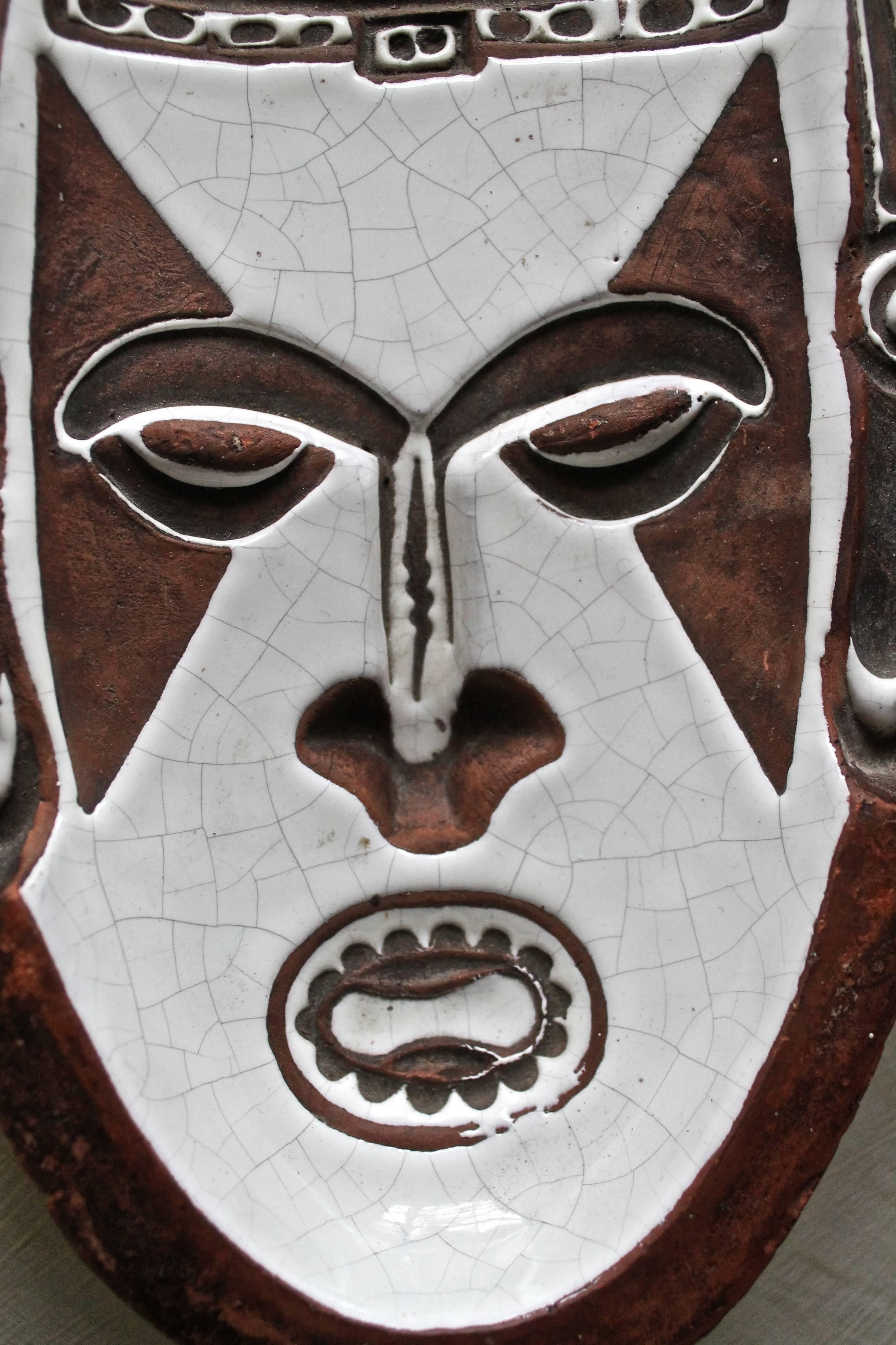 A low intaglio relief of a tribal mask, white crackle glaze on brown fired clay. Remnants of painted signature on bottom.