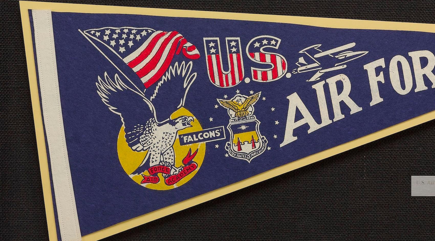 This vintage Air Force Academy pennant dates to the 1960s. The felt pennant has a blue background with a white headband. Decorated with the U.S. Flag pattern, the pennant has “U.S.” written just above the bold white “Air Force” with a depiction of