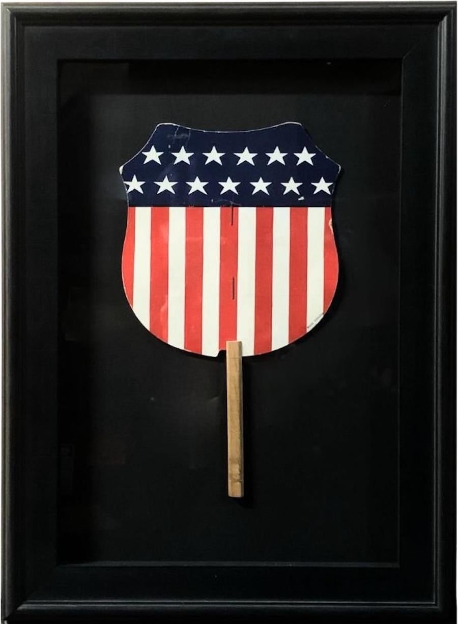 Presented is a U.S. shield patriotic printed paddle fan from 1918. The blue chief is printed with thirteen white stars. Below, thirteen vertical pales are printed in alternating red and white. This fan is attached to its original wooden stick and