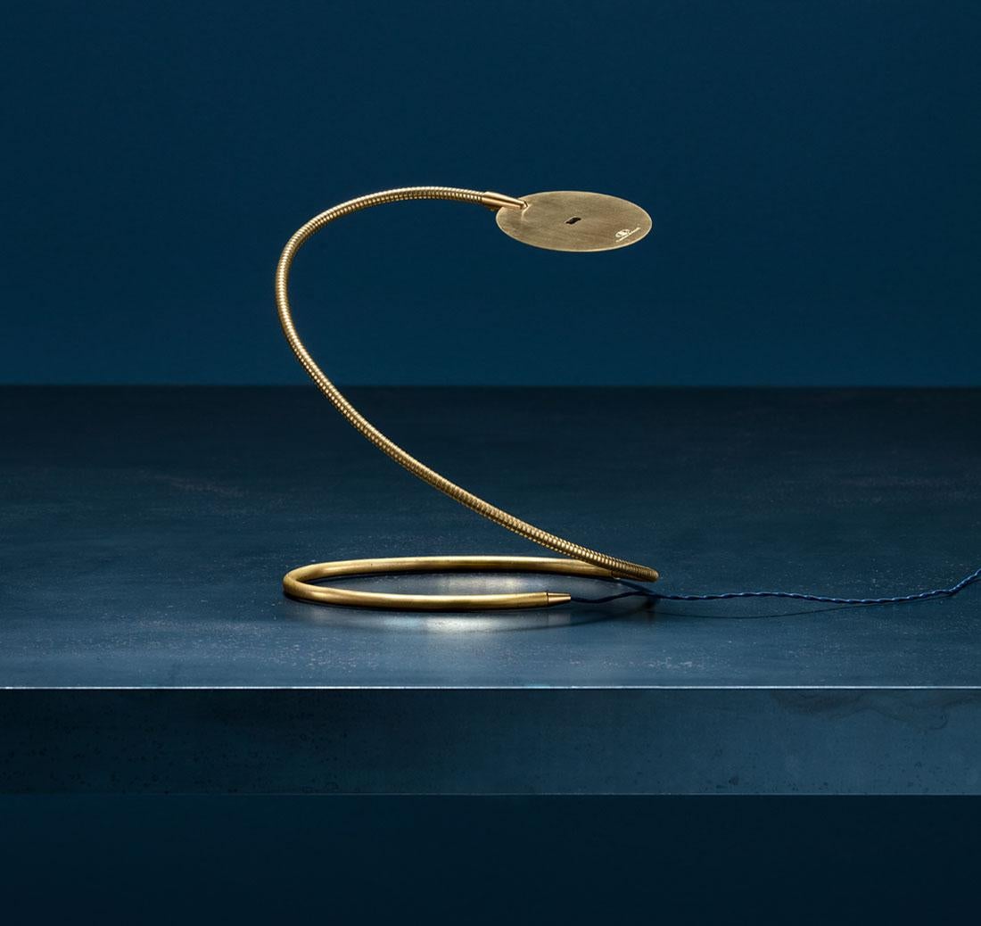 The Herem table lamp, crafted entirely from natural brass, embodies a harmonious balance of sculptural solidity and flexible movement. Its design combines a rigid base with a supple, adjustable arm that holds a slender oval shade. This gracefully
