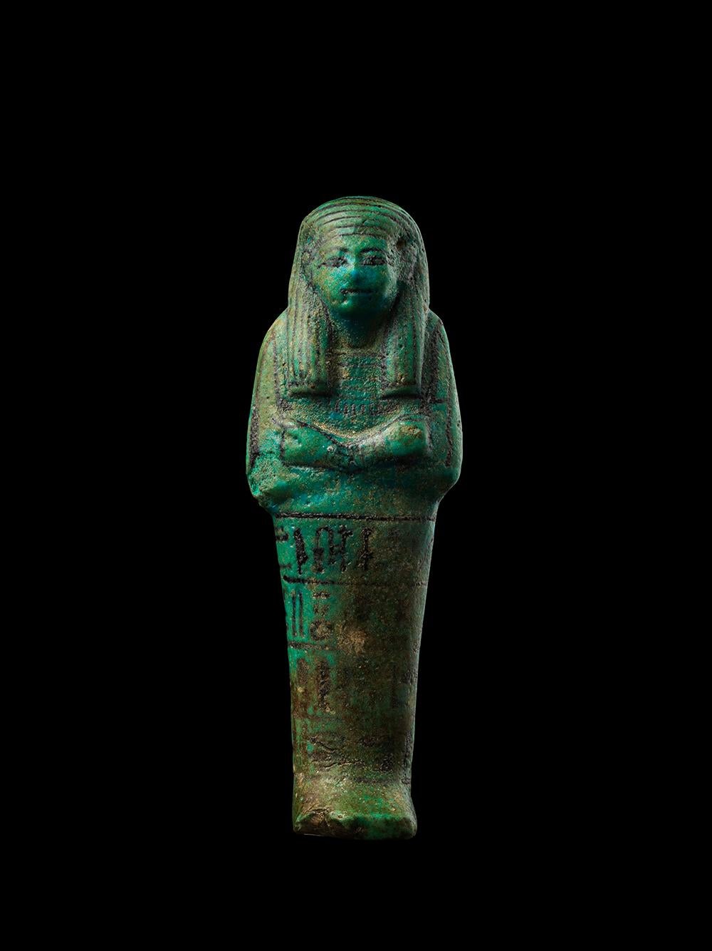 A mummiform ushabti in blue faience. Four registers of hieroglyphic characters are painted around the lower half of the ushabti, with a vertical column of hieroglyphs in the centre of the back. The inscription dedicates the ushabti to the royal