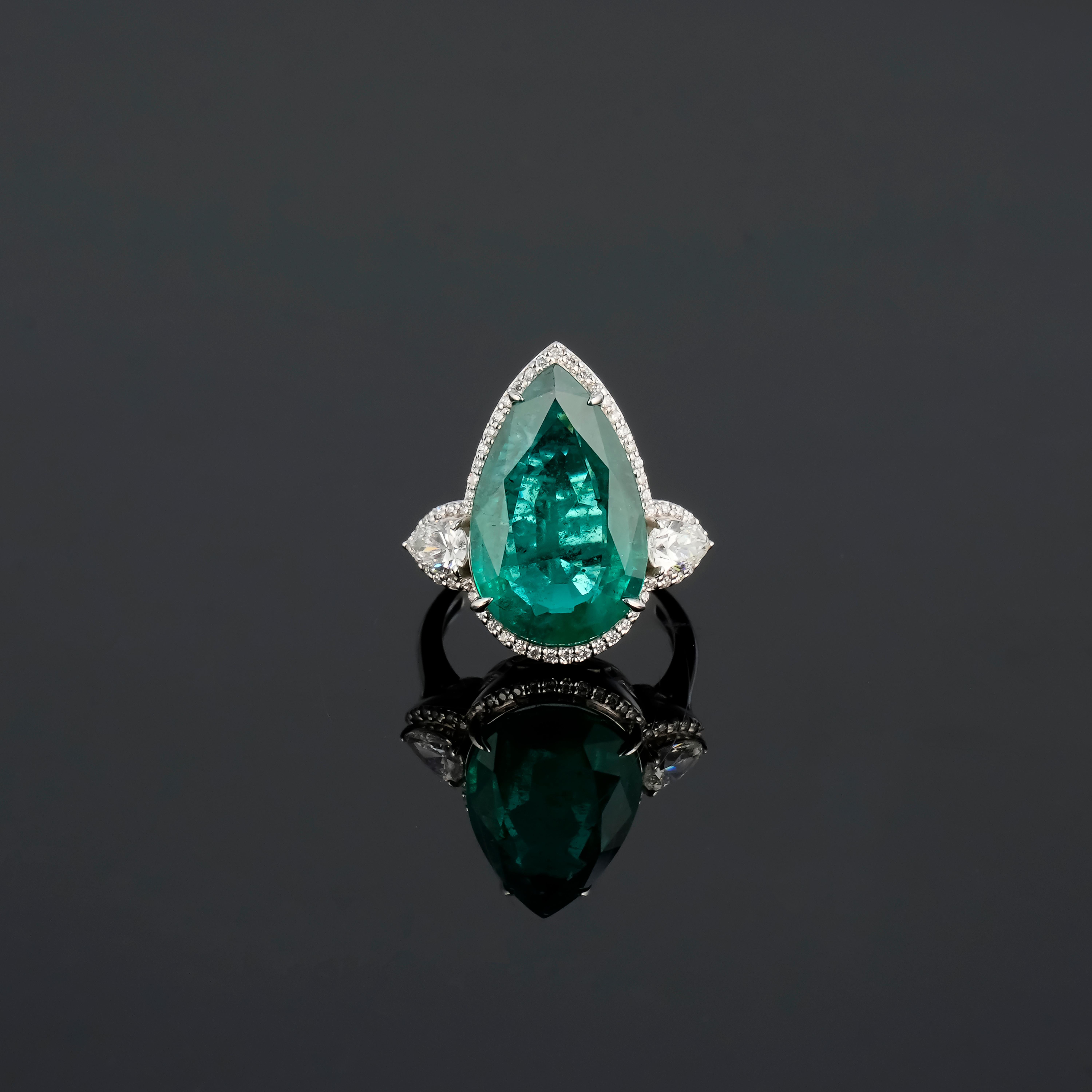Make a statement in this stunning 9.37 carat pear shaped Zambian Emerald center stone cocktail ring, adorned with 0.62 carat pear shape Diamond side stones, and 0.22 carat round Diamonds. The Emerald is transparent, with a beautiful vivid green