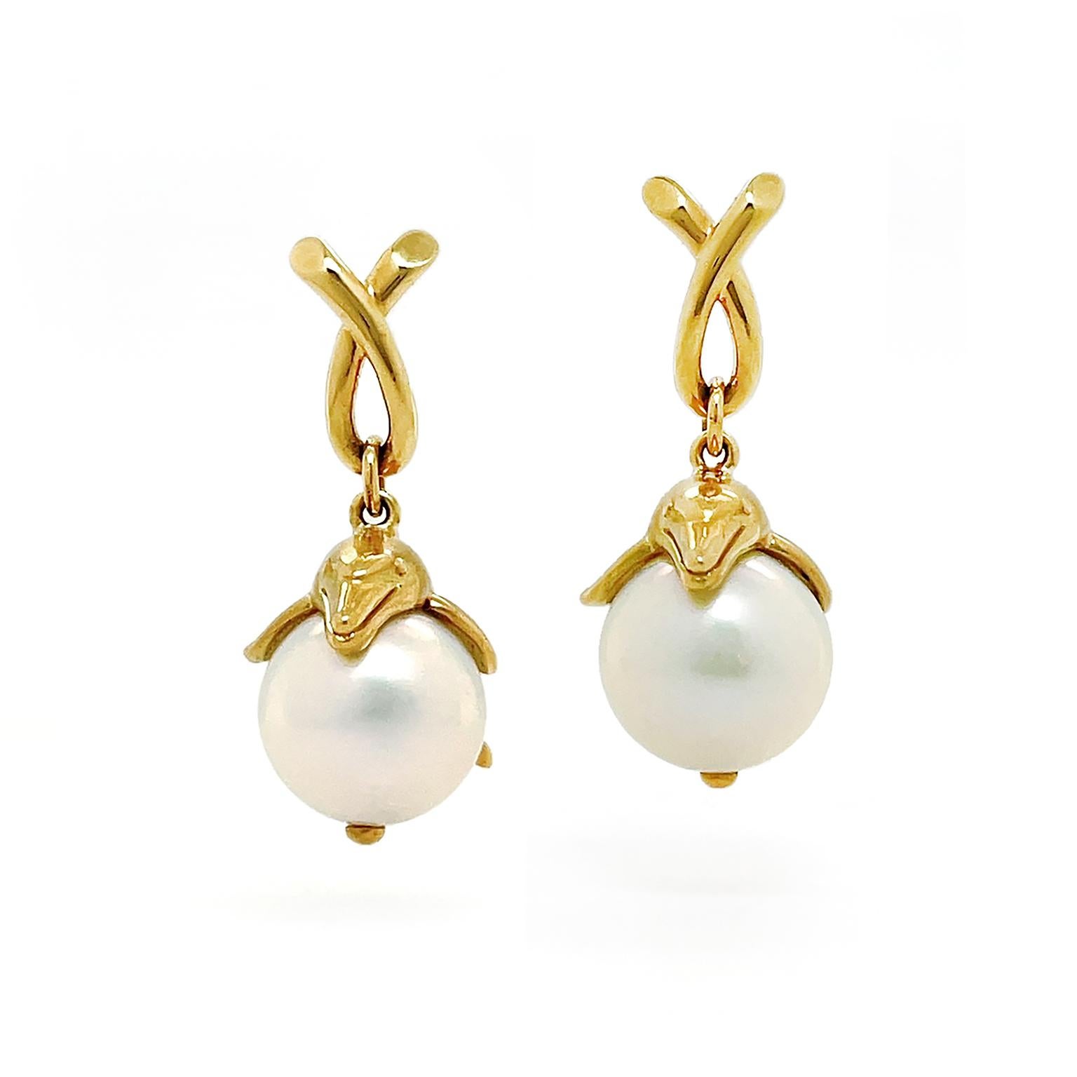 Dolphins produce a fanciful interpretation of pearl earrings. 18k yellow-gold dolphins drop from an overlapped gold loop. Detailing of the mammal includes its eyes and curved mouth. The position of the dolphin gives the illusion of leaping in midair