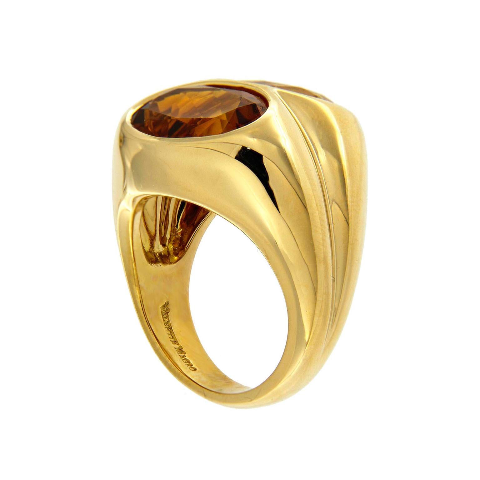 Two bands merge into a single ring. The 18k yellow gold body features a broad shank with a groove along its center, resembling two pieces merging. On top are faceted bezel set jewels close enough to touch. One is a saturated Madeira citrine, and the