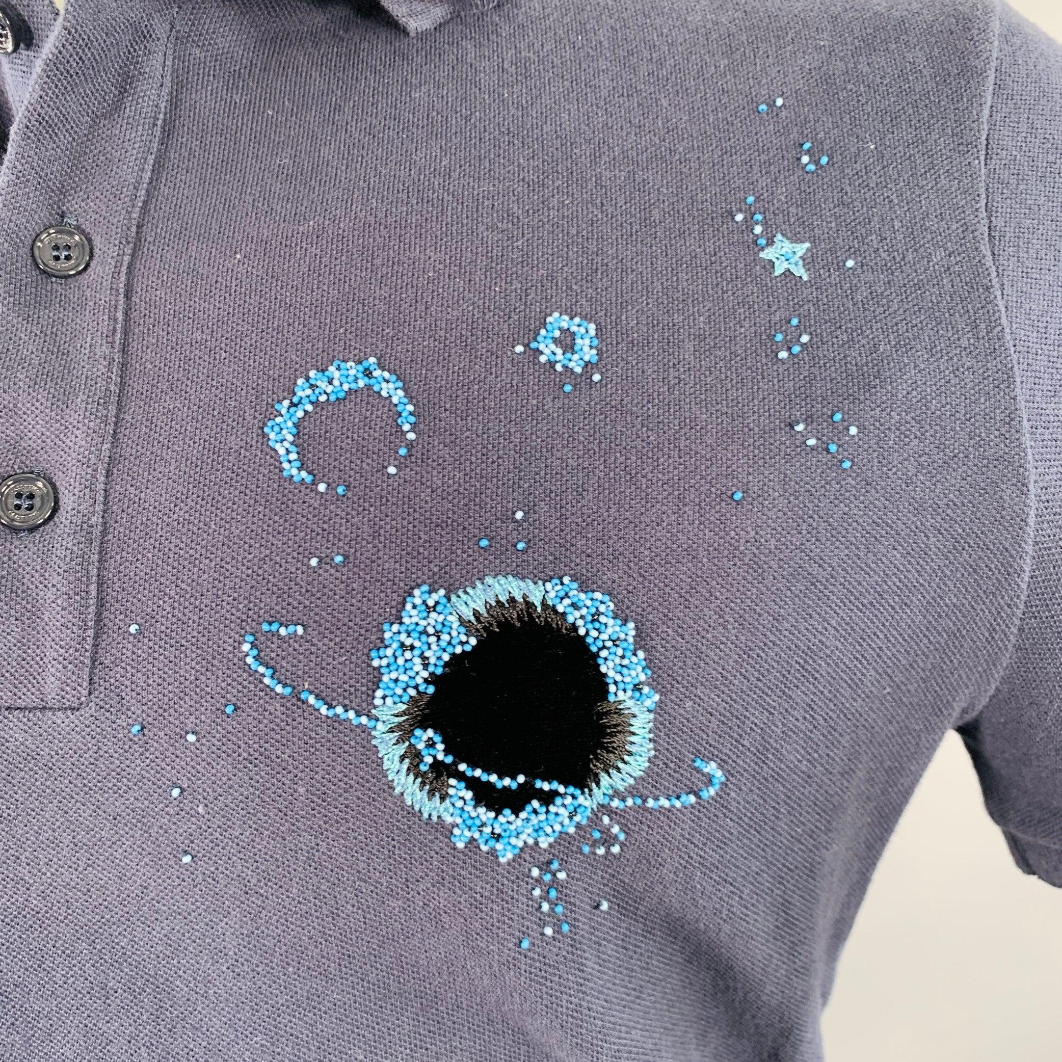 Polo VALENTINO
en piqué de coton bleu et marine, avec des planètes et des étoiles en perles bleues et des détails de planètes en velours noir, un texte en perles noires sur la manche gauche (qui dit : 