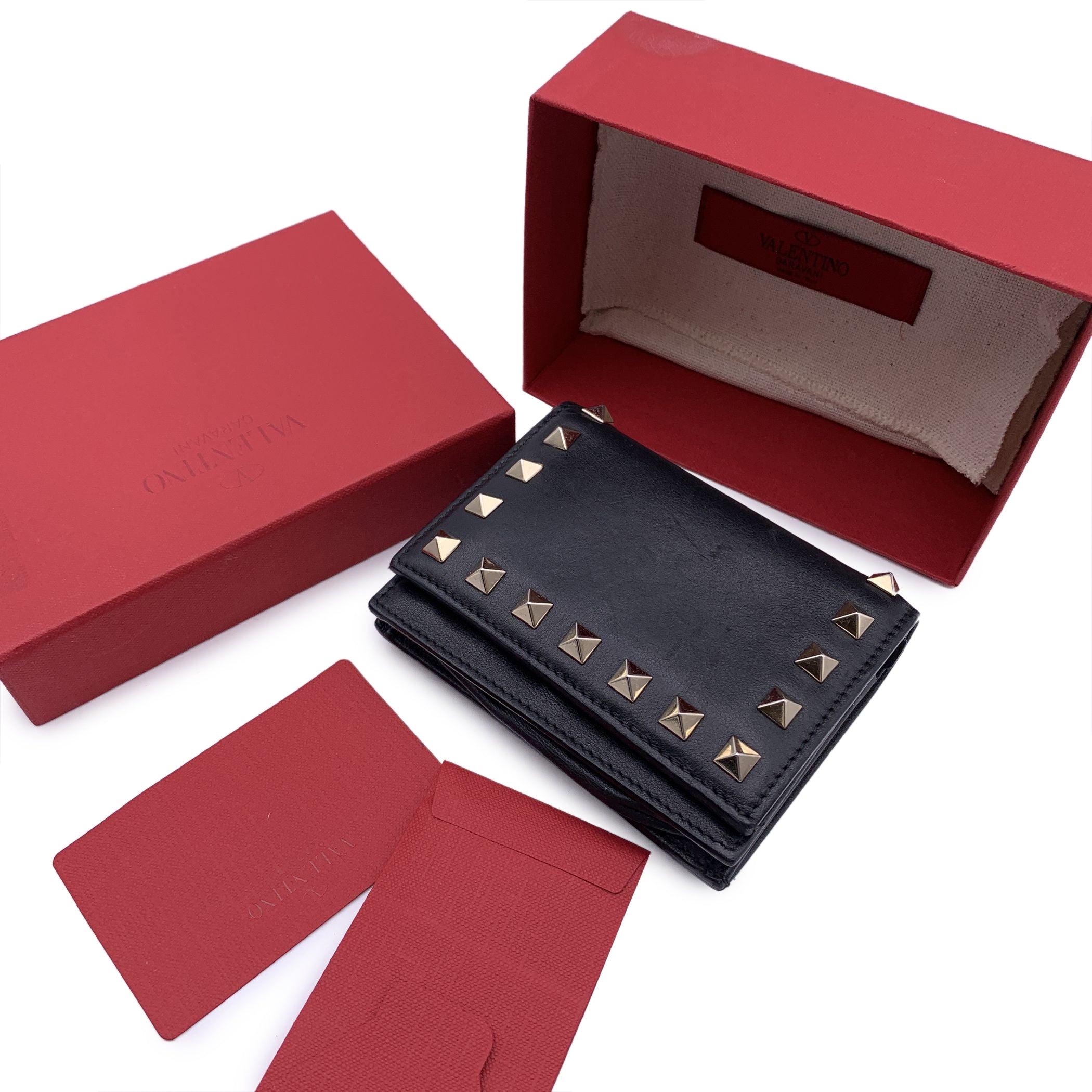 Valentino Garavani compact small compact wallet in black leather. Light gold metal pyramid studs detsialing. Logo embossed on leather on the back. Flap with button closure. Inside it has 5 credit card slots, 1 zip compsrtmante for coins and 1 bill