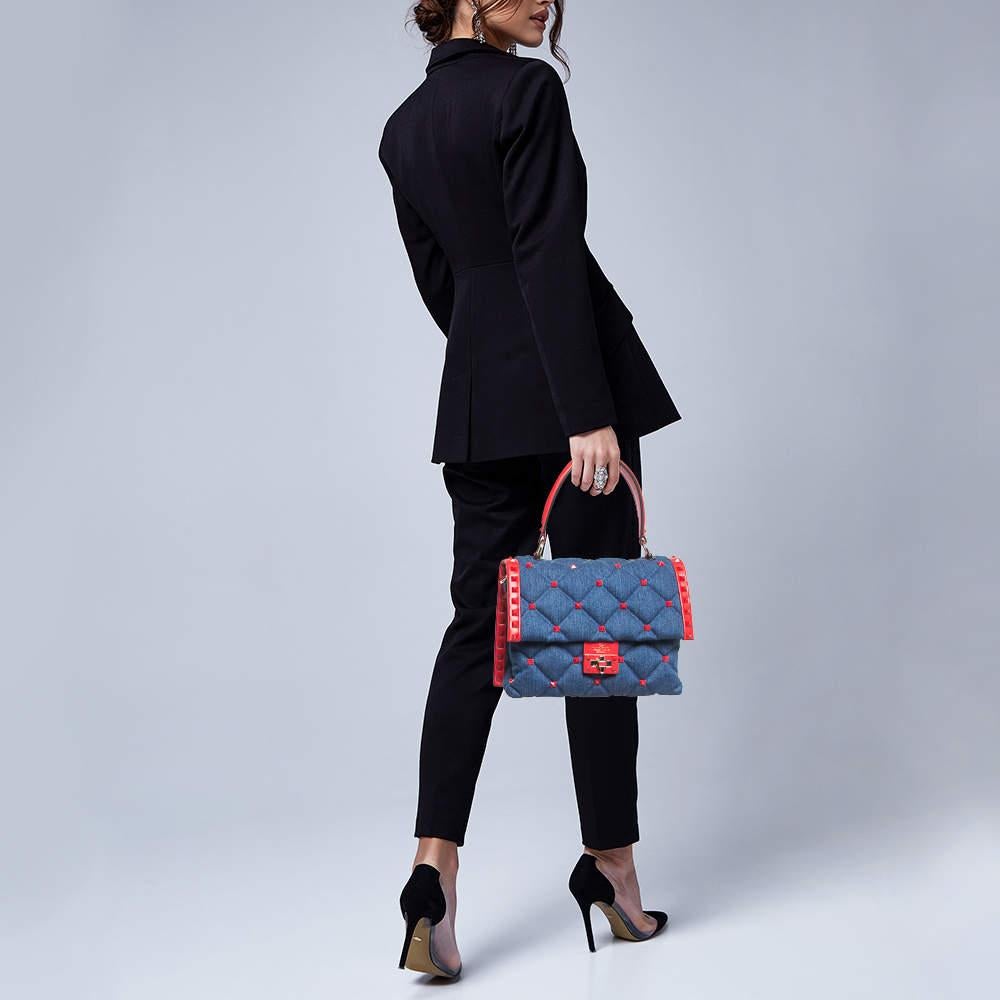 The Valentino Candystud Top Handle Bag is a stunning fusion of denim and supple leather, exuding effortless elegance. Its vibrant hues of blue and red harmonize seamlessly, adorned with the iconic Candystud embellishments. With its top handle