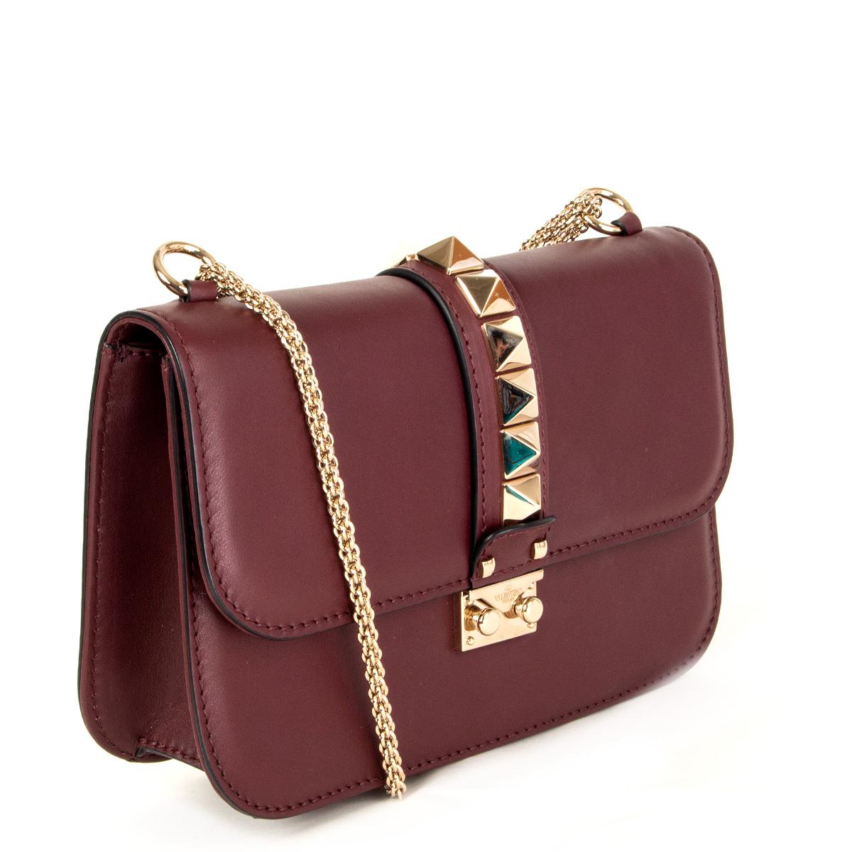 100% authentic Valentino Rockstud Glam Lock flap bag in burgundy calfskin features edgy, oversized pyramid studs in light gold-tone. A chain-link pull-through strap can be used as a shoulder strap, worn across the body, or removed to carry this bag