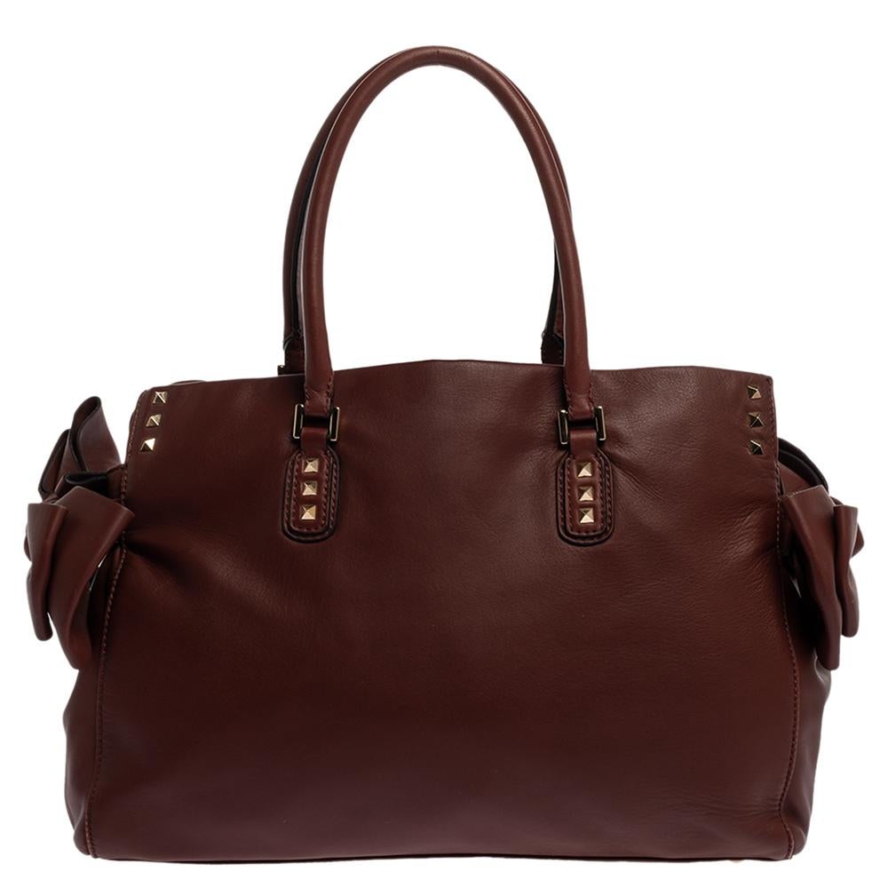 This stylish bow tote comes from the house of Valentino. Crafted meticulously in Italy, it is made from soft leather and comes in a lovely shade of burgundy. It is styled with dual handles, bow detailing on the sides, Rockstuds, gold-tone hardware