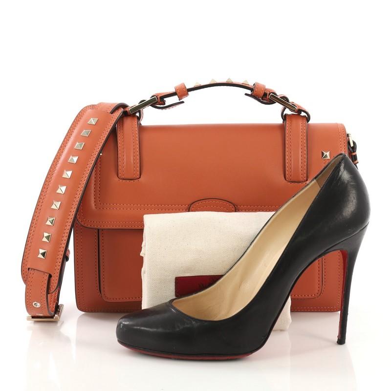 This Valentino Cabana Top Handle Flap Bag Leather Medium, crafted from orange leather, features a studded leather top handle, slip pocket under flap, and gold-tone hardware. Its push-lock closure opens to an orange leather interior with two open
