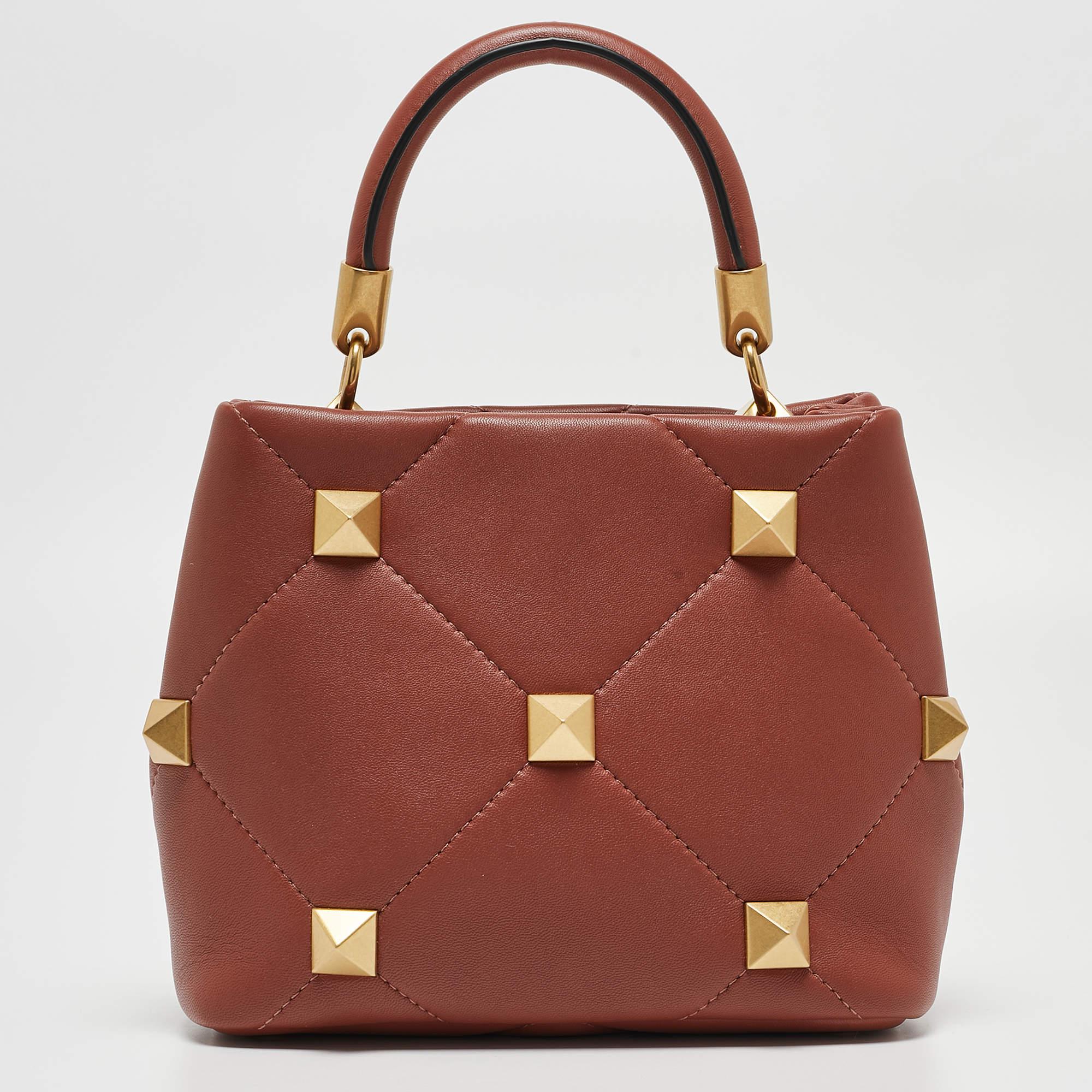 The Valentino top handle bag epitomizes timeless elegance. Crafted from luxurious chestnut brown leather, and adorned with exquisite Roman stud detailing, this sophisticated accessory seamlessly blends classic design with modern allure. Elevate your