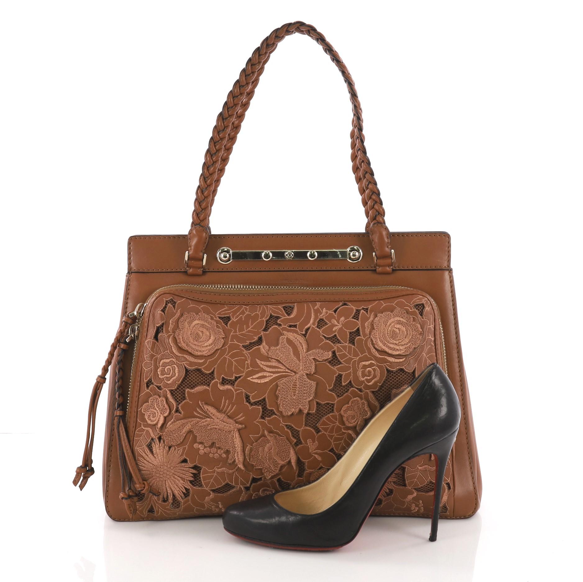 This Valentino Demetra Tote Leather Lace, crafted in brown leather, features braided leather straps, a large front zip-around pocket with braided zipper pulls and cutwork lace front panel, protective base feet and gold-tone hardware. Its push-lock