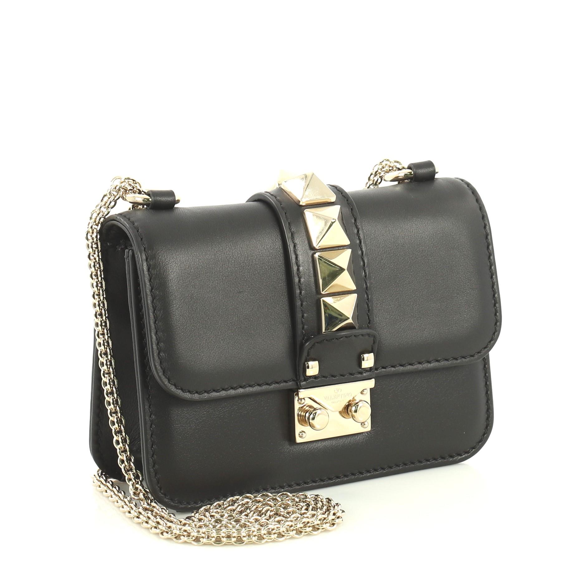 This Valentino Glam Lock Shoulder Bag Leather Mini, crafted from black leather, features a long detachable chain strap, pyramid studs at the center, and gold-tone hardware. Its push-lock closure opens to a black fabric interior with slip pocket.