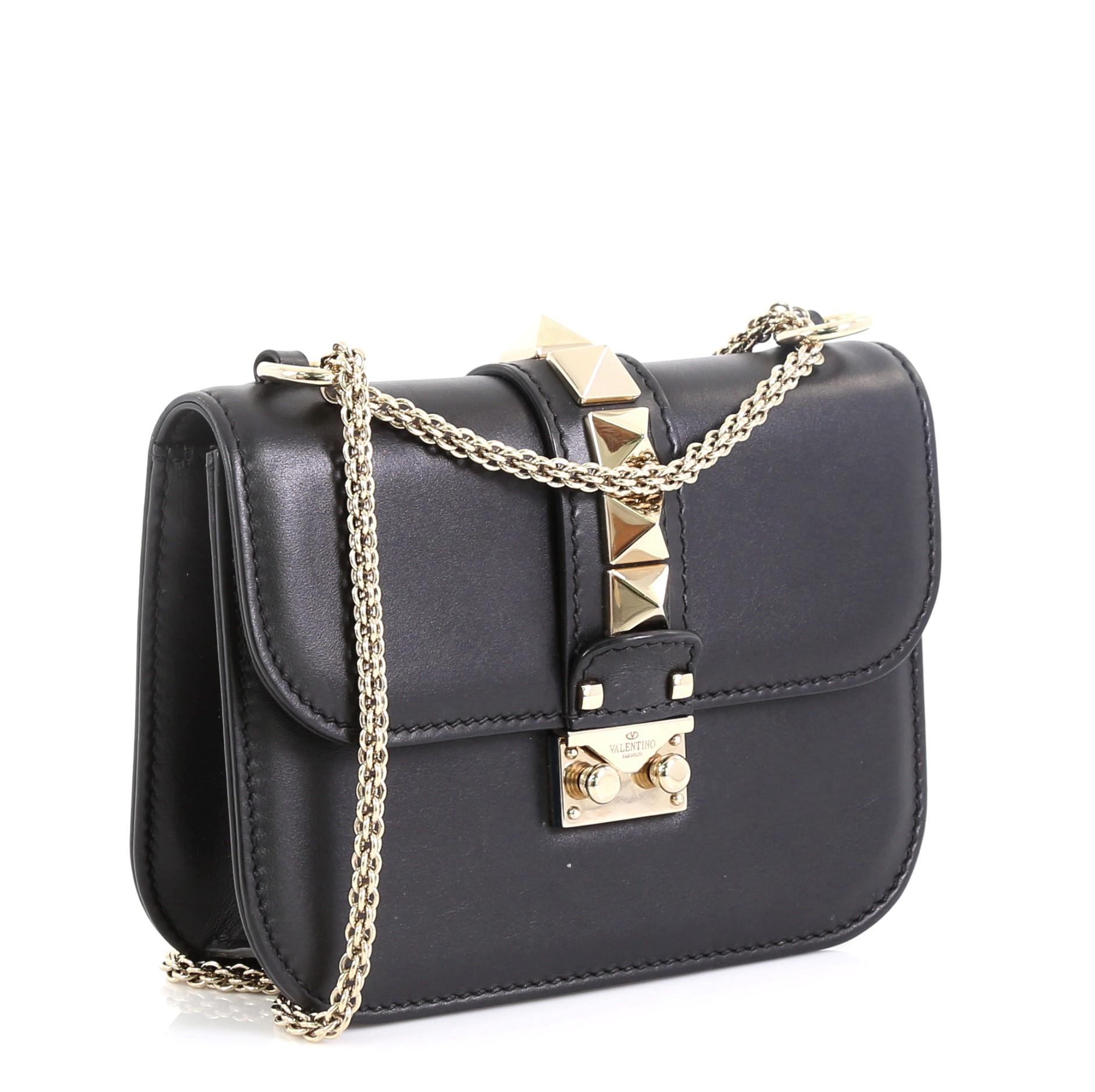 This Valentino Glam Lock Shoulder Bag Leather Small, crafted in black leather, features chain link strap, pyramid studs, and gold-tone hardware. Its push-lock closure opens to a black fabric interior with zip and slip pockets. 

Estimated Retail