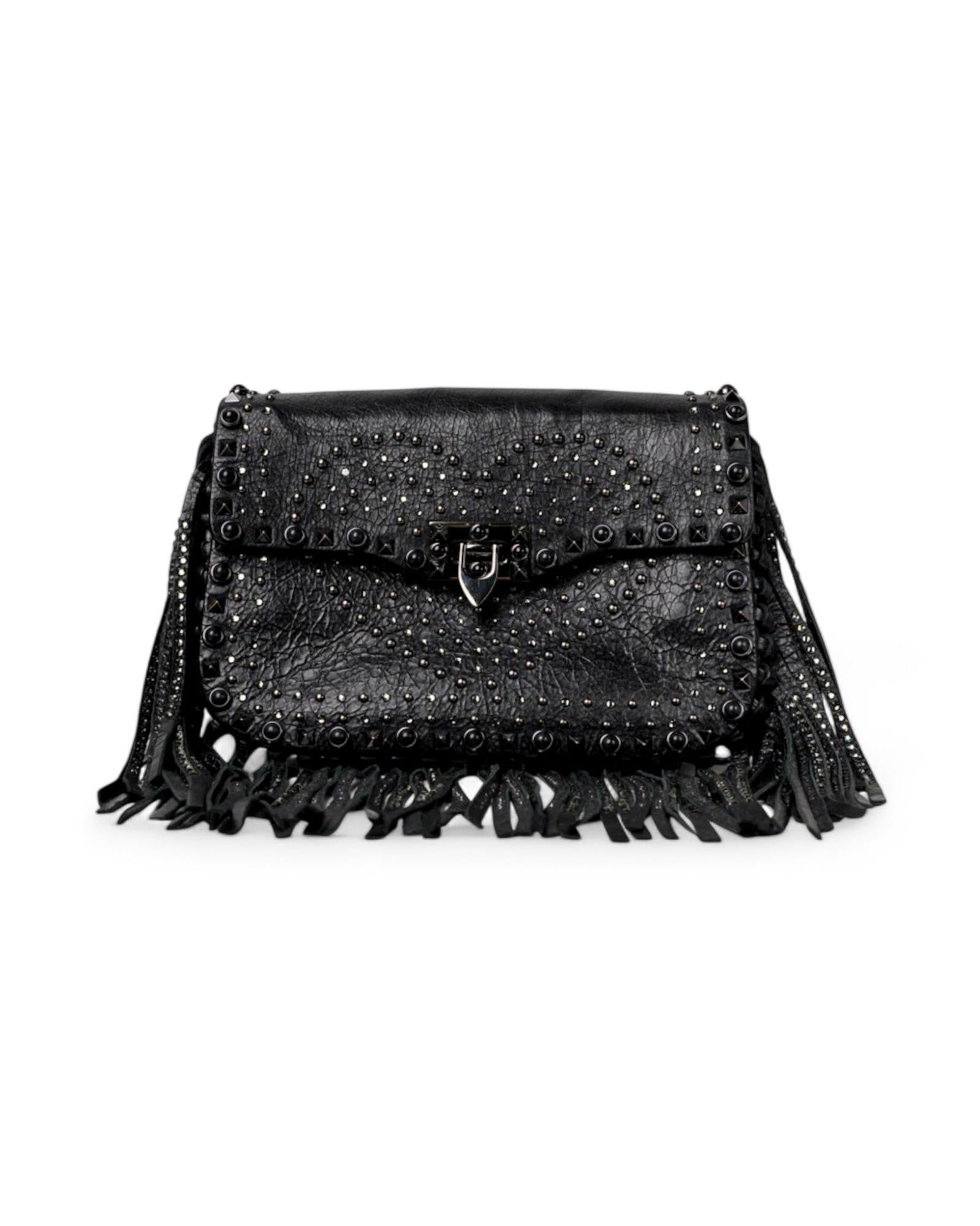 Bolso de firma Valentino, línea Rockstud, modelo Guitar en talla Small. Confeccionado en suave piel negra, adornado con tachuelas y acabados bruñidos, este bolso combina audacia y sofisticación. Presenta una solapa frontal con cierre entrelazado, el