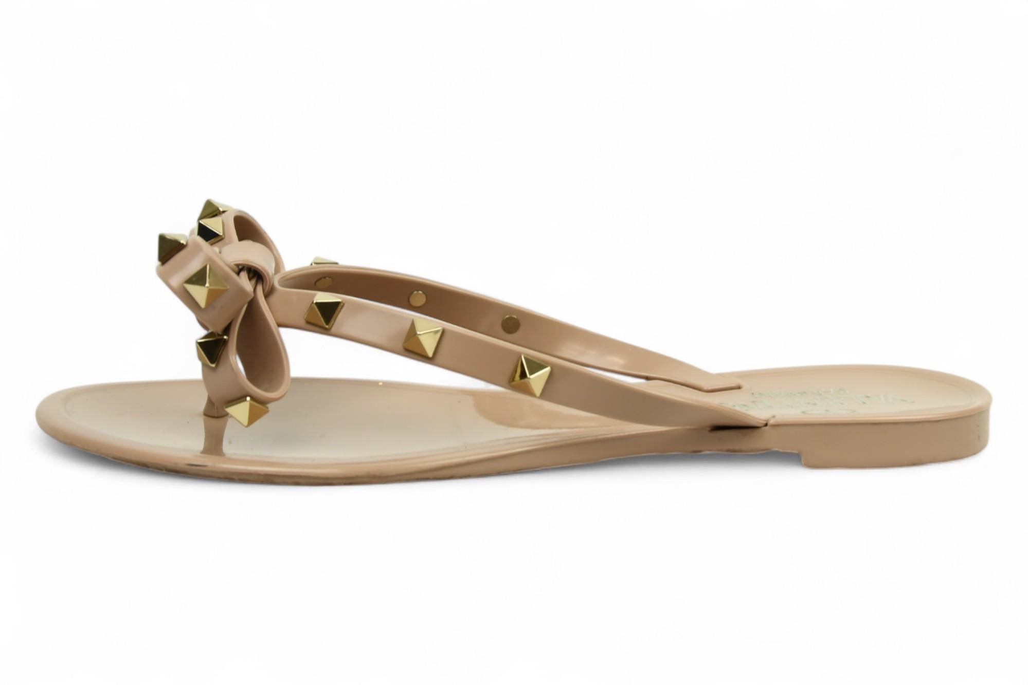 L'allegria incontra l'eleganza in questi sandali Valentino Garavani Rockstud, realizzati in gelatina lucida color nude con un fiocco e borchie a piramide color oro. Leggeri e resistenti all'acqua, questi sandali infradito combinano l'iconico fascino