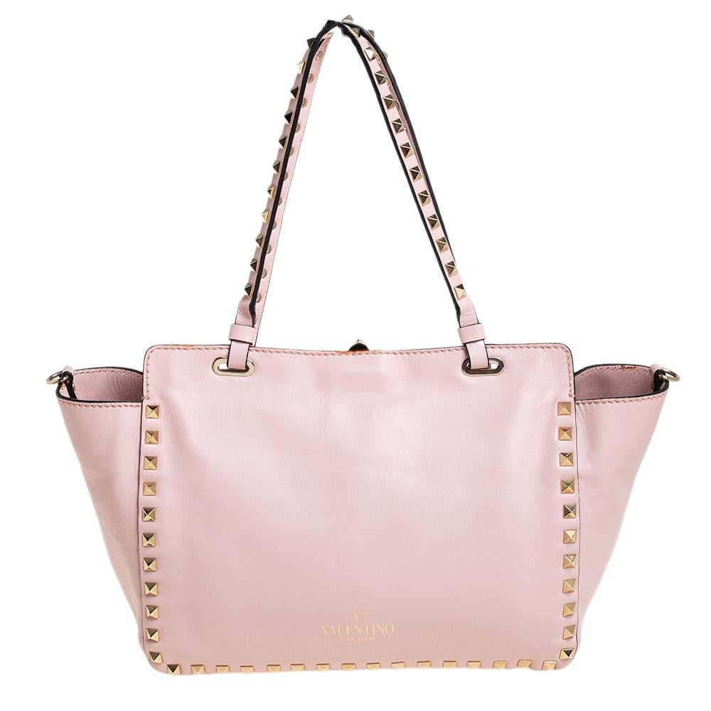La lujosa casa de moda italiana Valentino es famosa por sus diseños clásicos impregnados de su propio toque moderno y detalles Rockstud, como muestra este bolso tote trapecio. Fabricada en piel rosa, tiene asas superiores y una correa para el hombro
