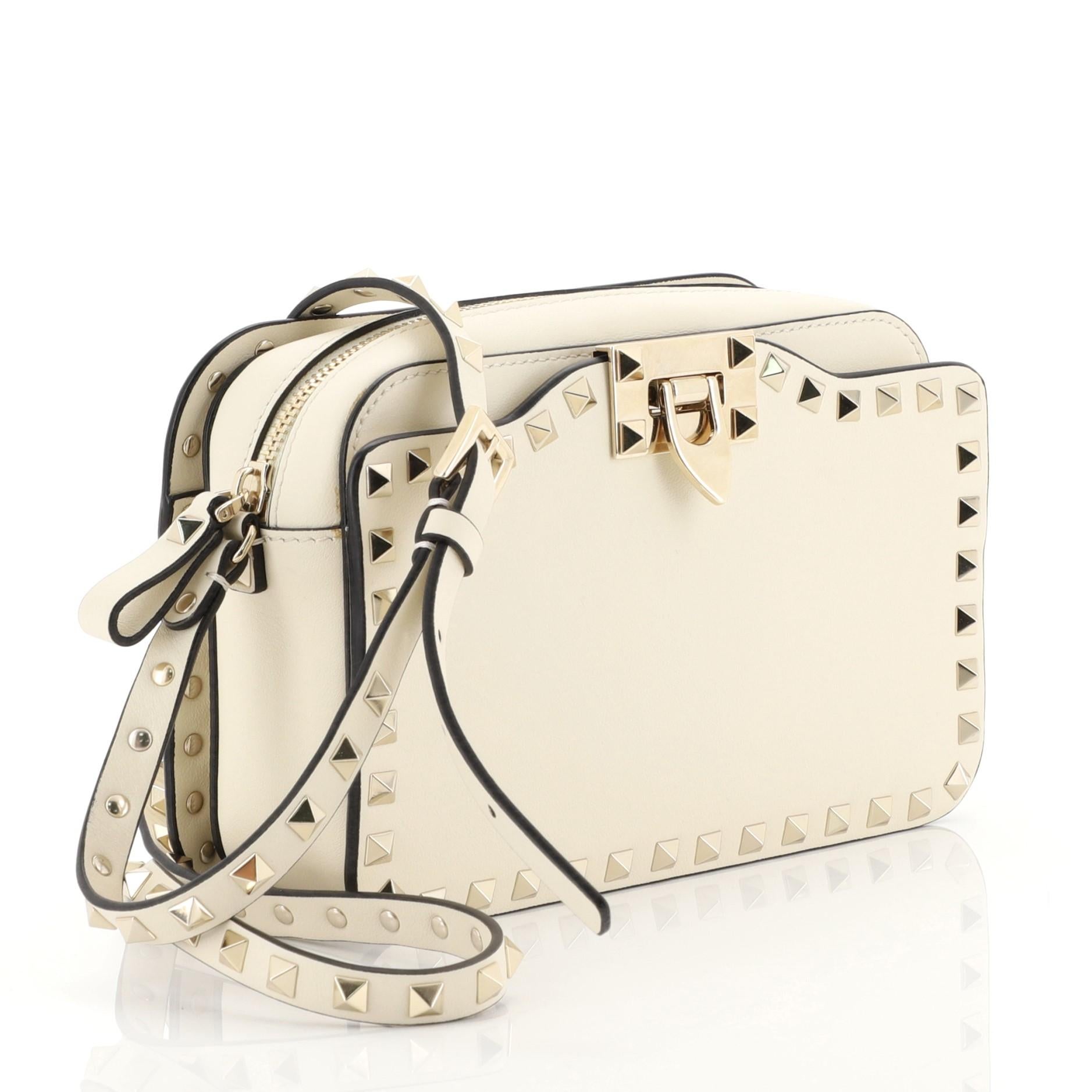 This Valentino Rockstud Camera Crossbody Bag Leather, crafted from white leather, features an adjustable strap, pyramid rockstud detailing, and gold-tone hardware. Its zip closure opens to a neutral fabric interior with slip pocket. 

Estimated